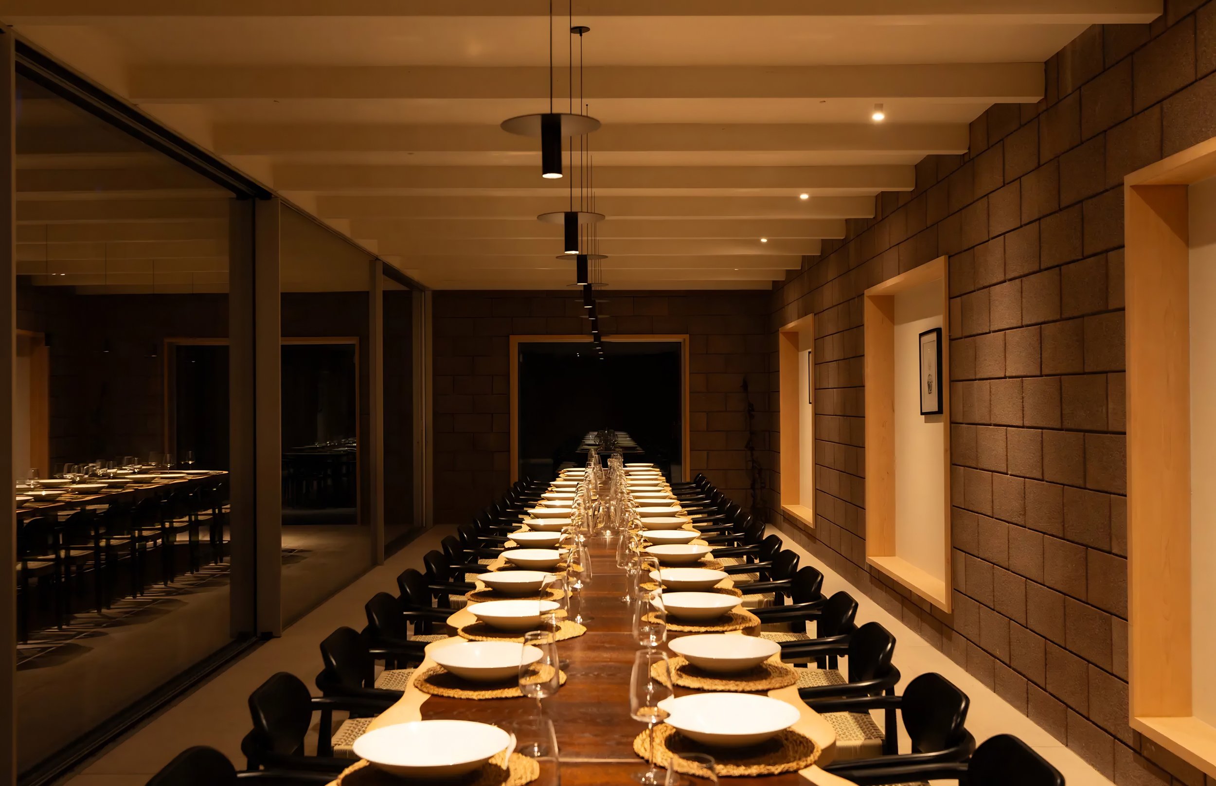 A long dining table set for a meal with white bowls, glasses, and woven placemats in a modern restaurant with warm lighting and brick walls.