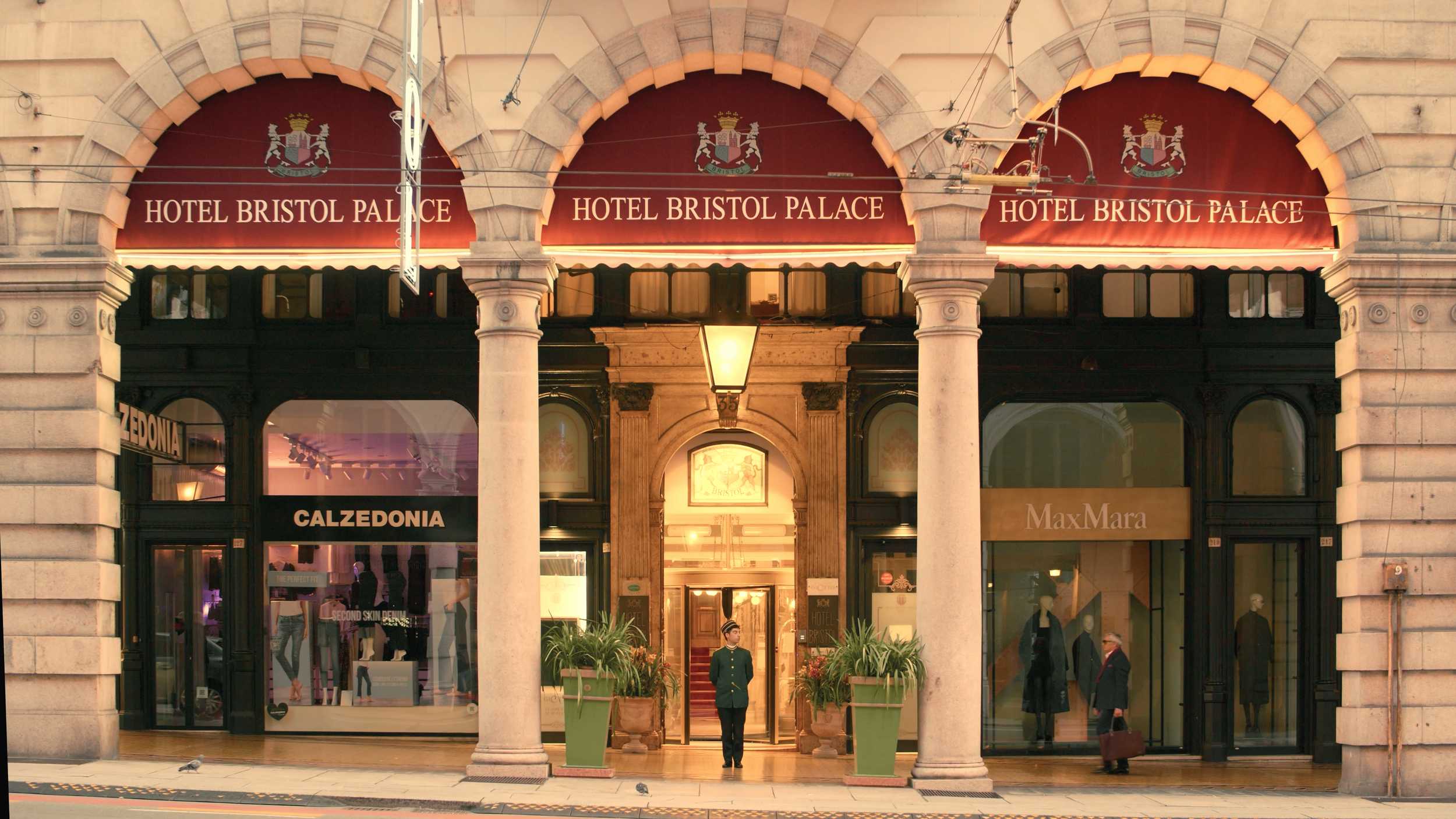  storefront of Hotel Bristol Palace with a red awning, arched windows, and stone columns, located on a city street with pedestrians and storefronts, including Calzedonia and MaxMara.