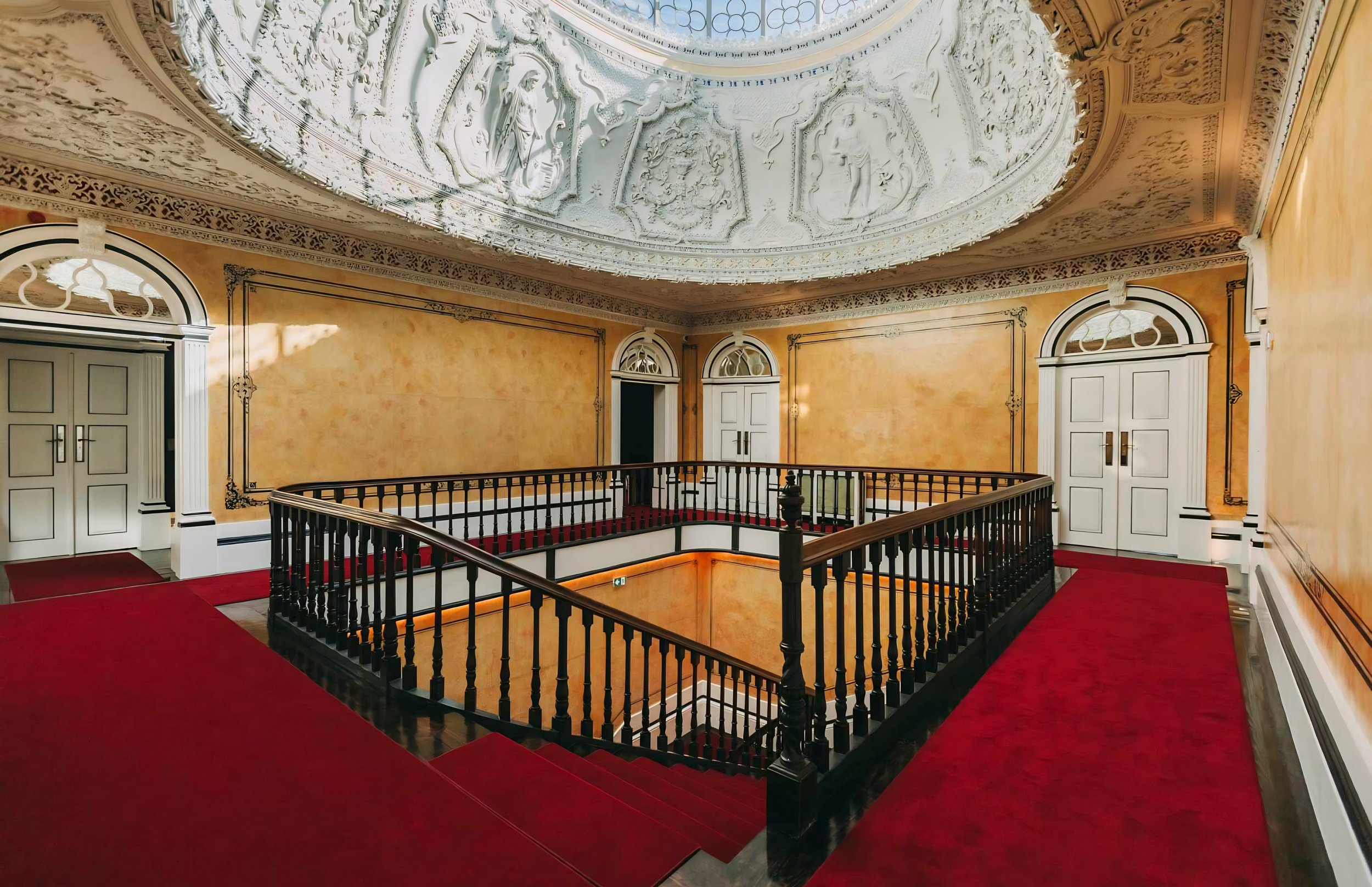 Interior of a building with ornate ceiling decorations, white double doors, and red carpeted stairs surrounding a black railing.