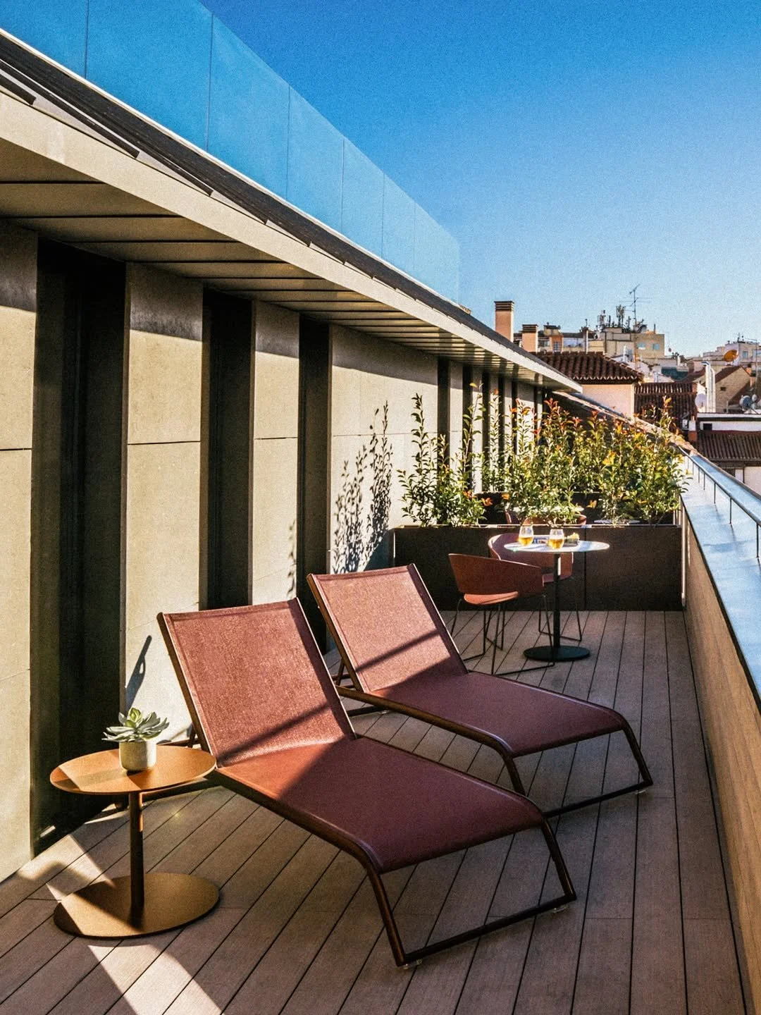 A rooftop terrace with two pink lounge chairs and small round tables, a row of potted plants, and city buildings in the background under a clear blue sky.