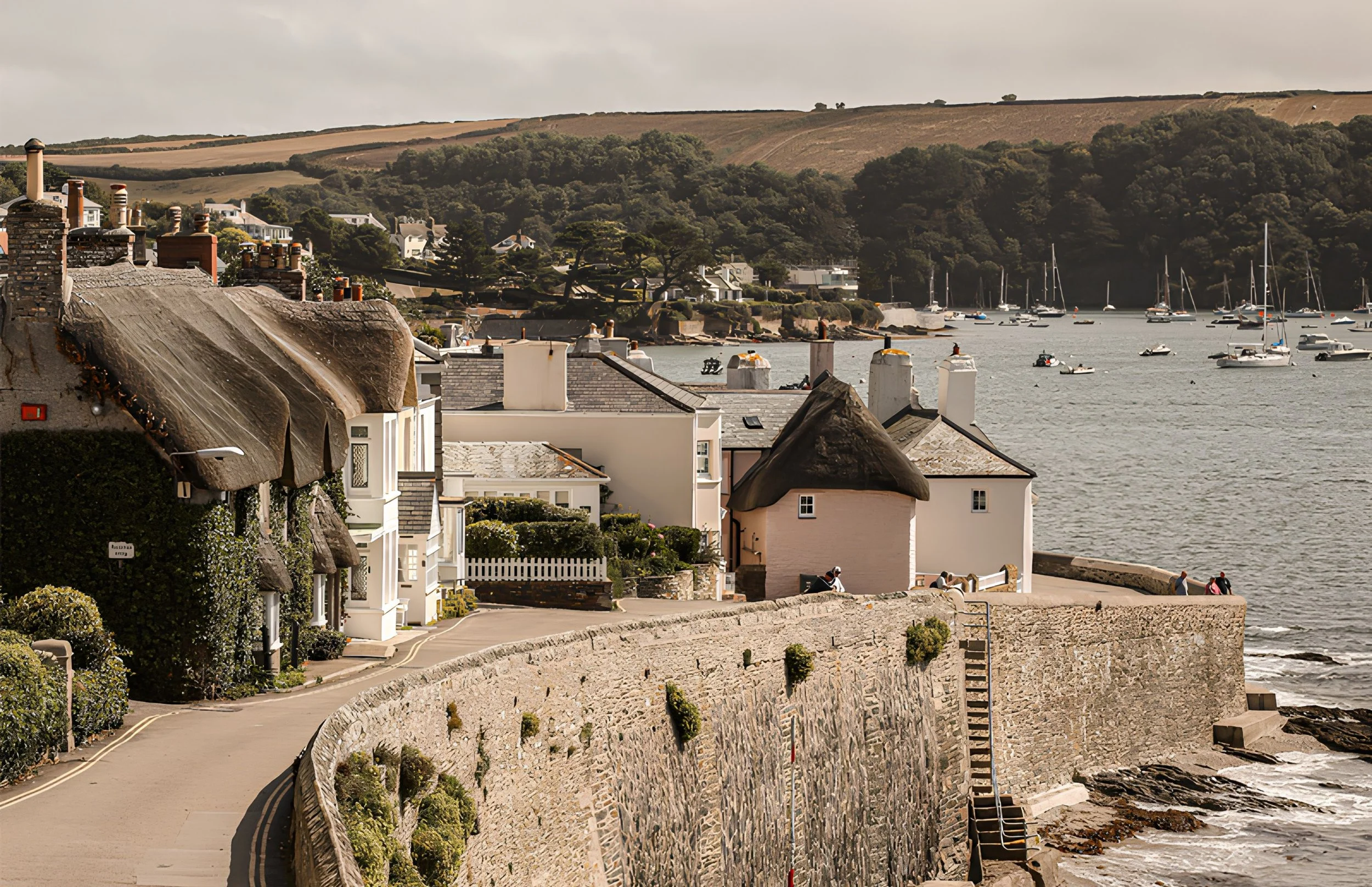 A coastal village with houses featuring thatched roofs along a stone seawall, overlooking a bay with sailboats anchored in calm water, hills in the background, and an overcast sky.