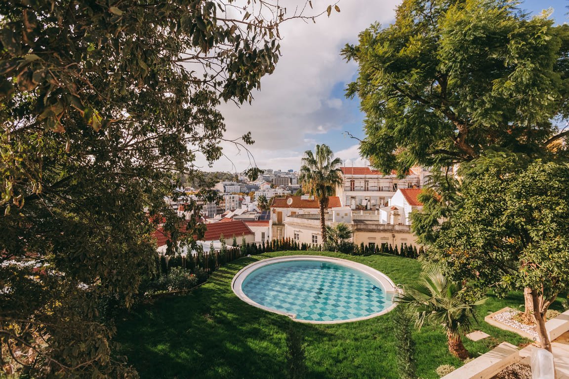 An aerial view of a backyard with a small, circular swimming pool surrounded by lush green grass and trees, overlooking a cityscape with various white buildings and red-tiled roofs.
