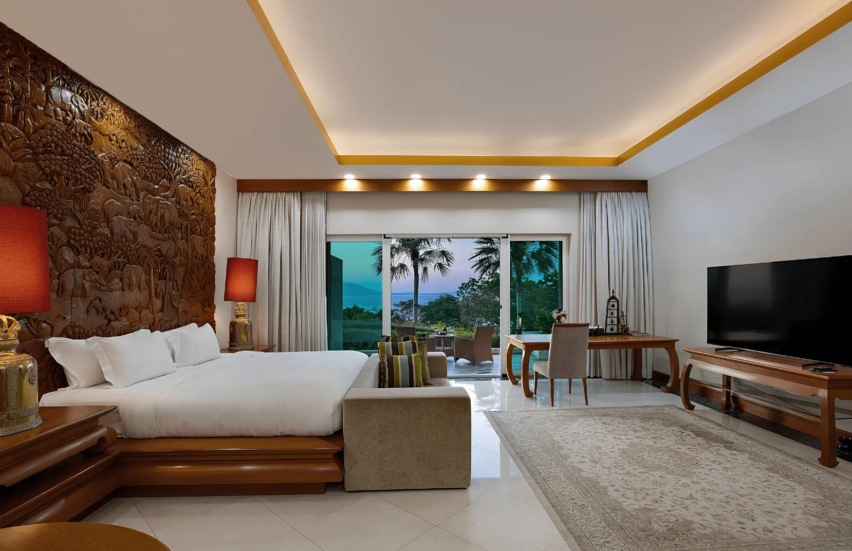 Elegant hotel room with a view of a patio, palm trees, and ocean through sliding glass doors, featuring a bed, side tables with lamps, a sitting area, a desk, and a large flat-screen TV.