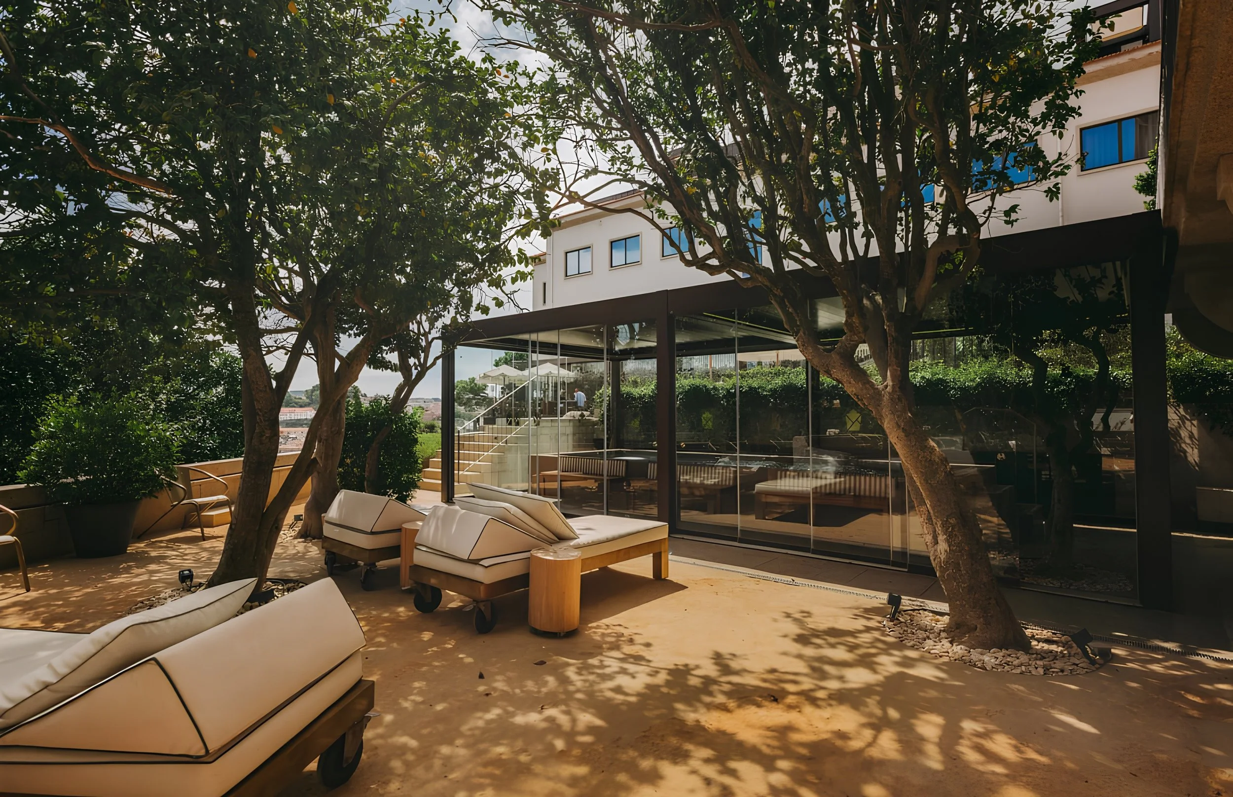 Outdoor patio area with lounge chairs, large trees providing shade, modern building in the background, glass windows reflecting the trees, and a staircase leading to an upper level.