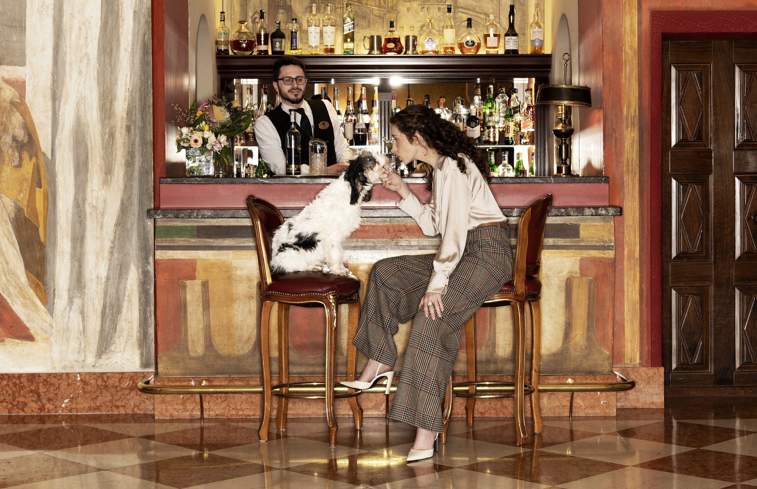 A woman with curly hair in plaid pants and white shoes sits at a bar, gently holding a small dog while leaning forward. Behind the bar, a bartender with glasses and a vest looks on. The bar is decorated with flowers, and various bottles of alcohol ar