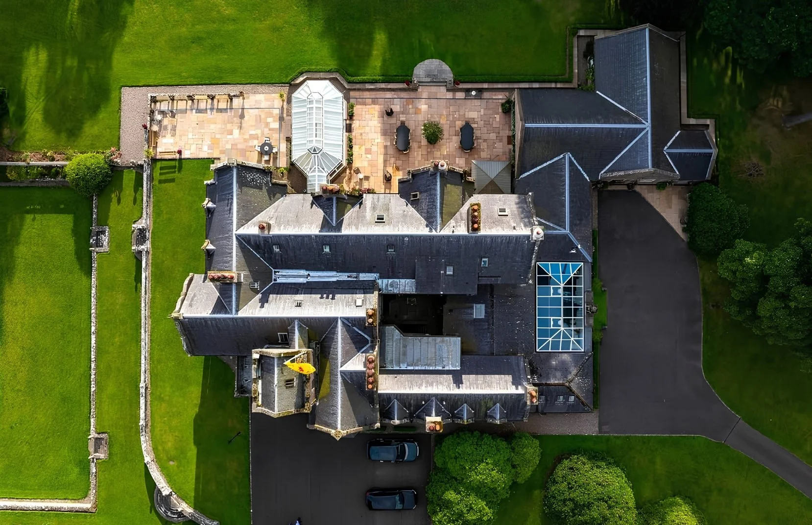 Aerial view of a large house with a black roof, surrounded by green lawns and trees, with a patio area and driveway visible.