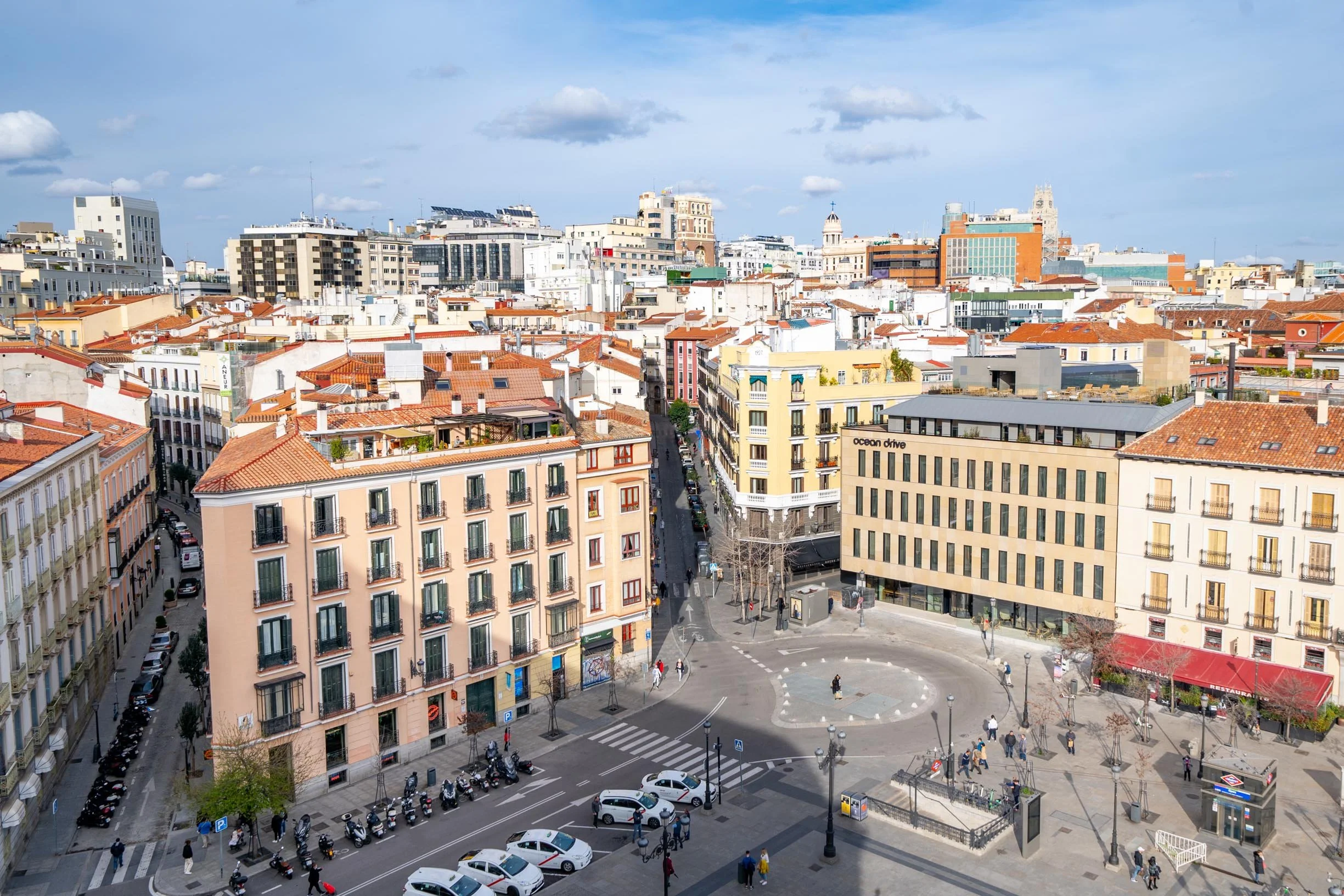 Cityscape view with multiple buildings, rooftops, and a street corner with parked motorcycles and cars, pedestrians walking on sidewalks, and a partly cloudy sky.