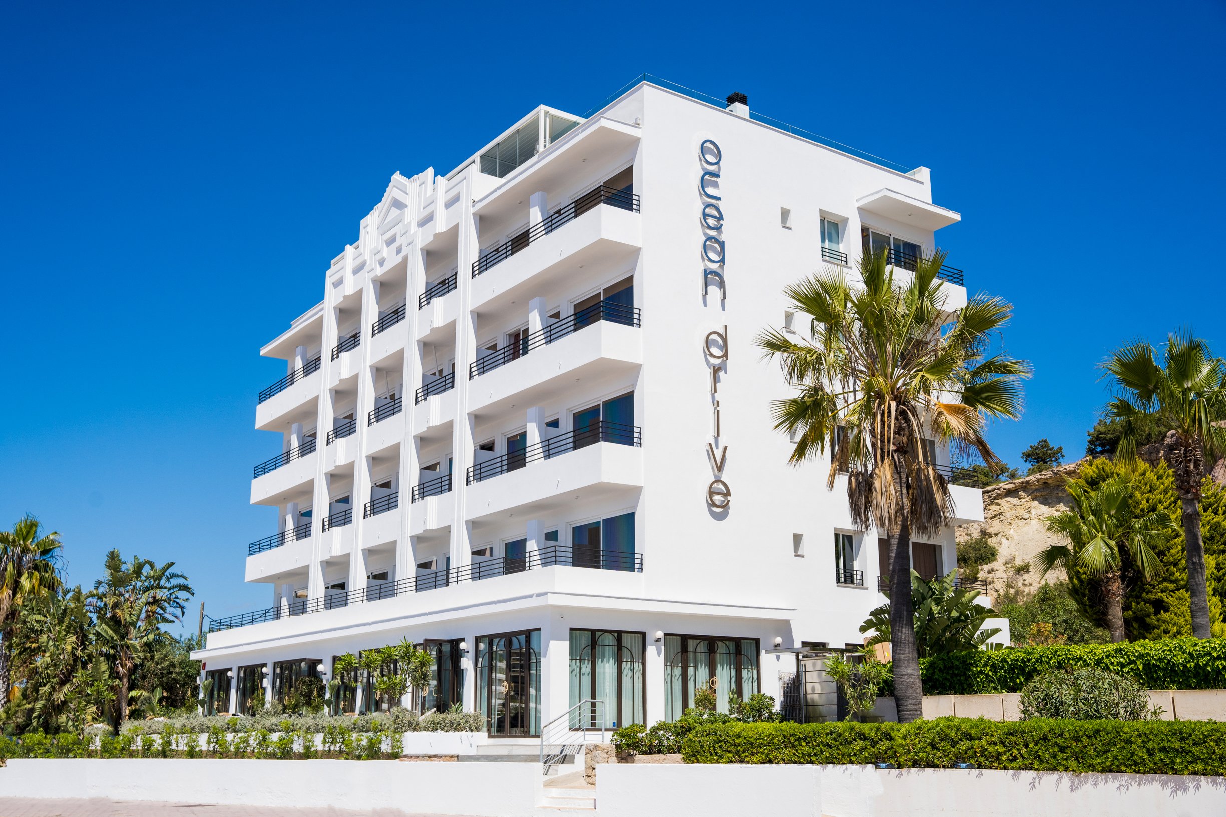 A modern white hotel building with multiple balconies, surrounded by palm trees and green plants, under a clear blue sky.