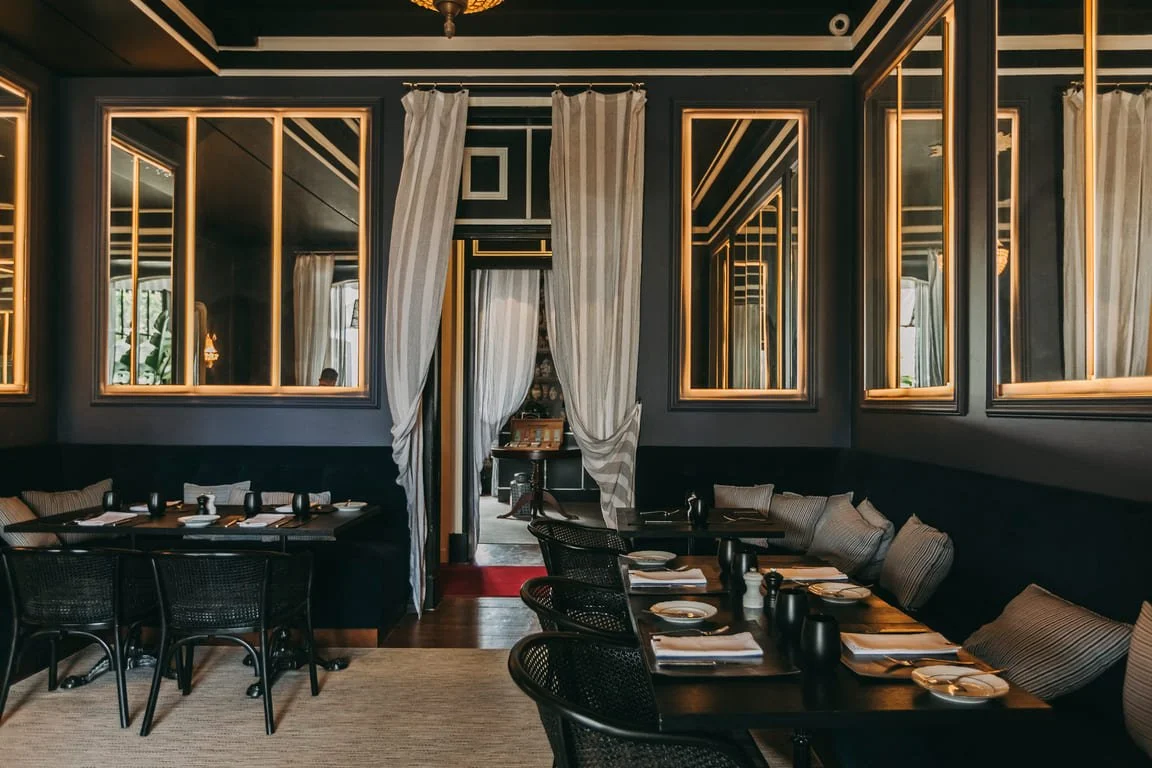 A stylish dining room featuring dark walls with gold accents, mirror panels, and striped curtains. The room includes two tables set with dishes, utensils, and black cups, surrounded by black chairs and a cushioned bench, with a doorway lined with flowing curtains leading to another room.