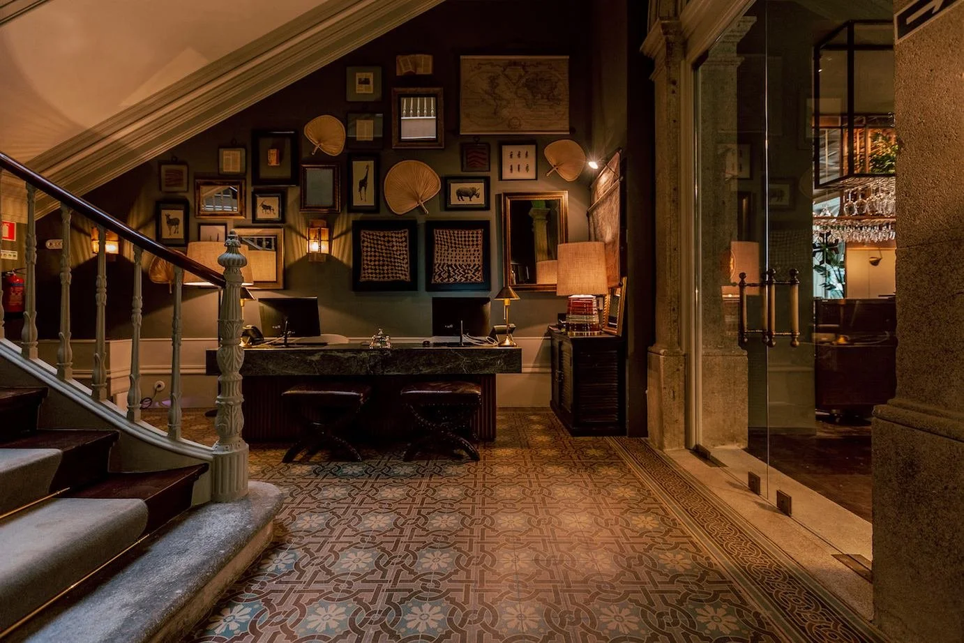 Hotel reception area with a dark green wall decorated with framed pictures, fans, and a world map. A stone desk with lamps and a computer, and a staircase with white railings leading up on the left. Glass doors on the right reveal a dimly lit lounge 