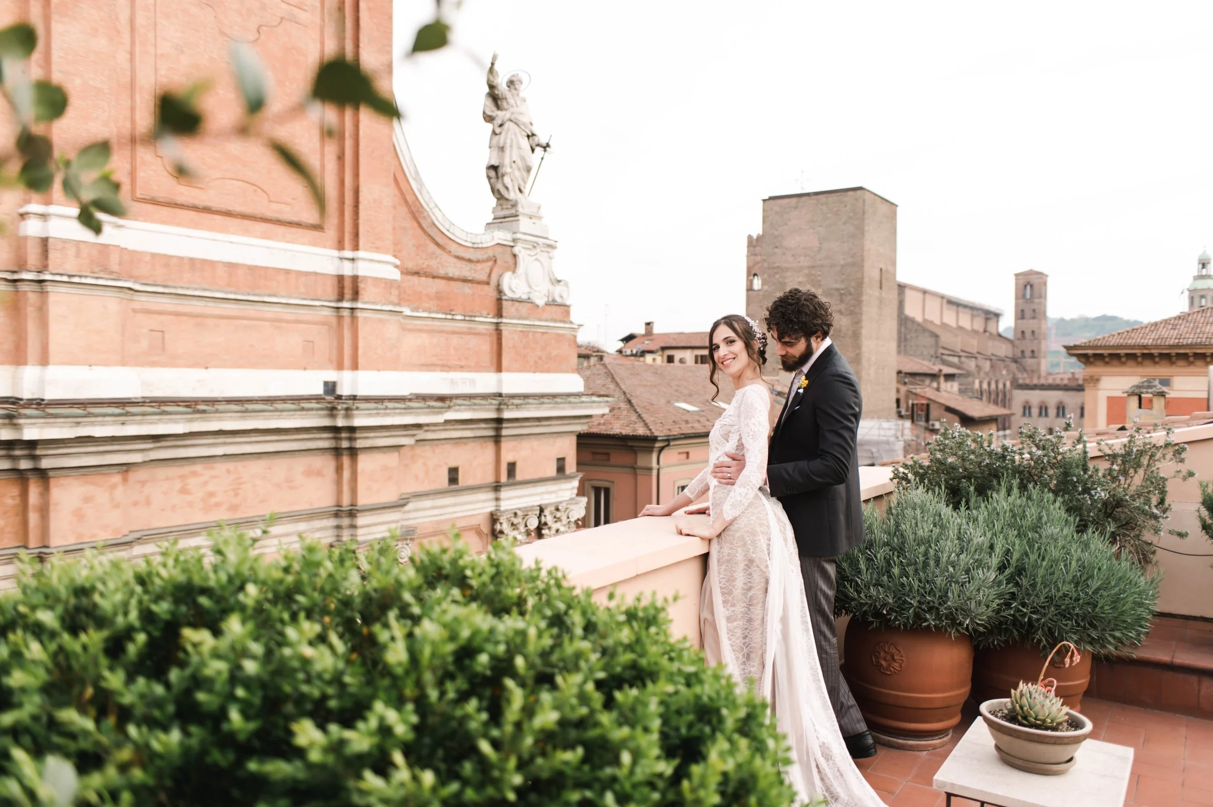 A bride and groom standing on a rooftop terrace with potted plants, with historical buildings and a statue in the background.