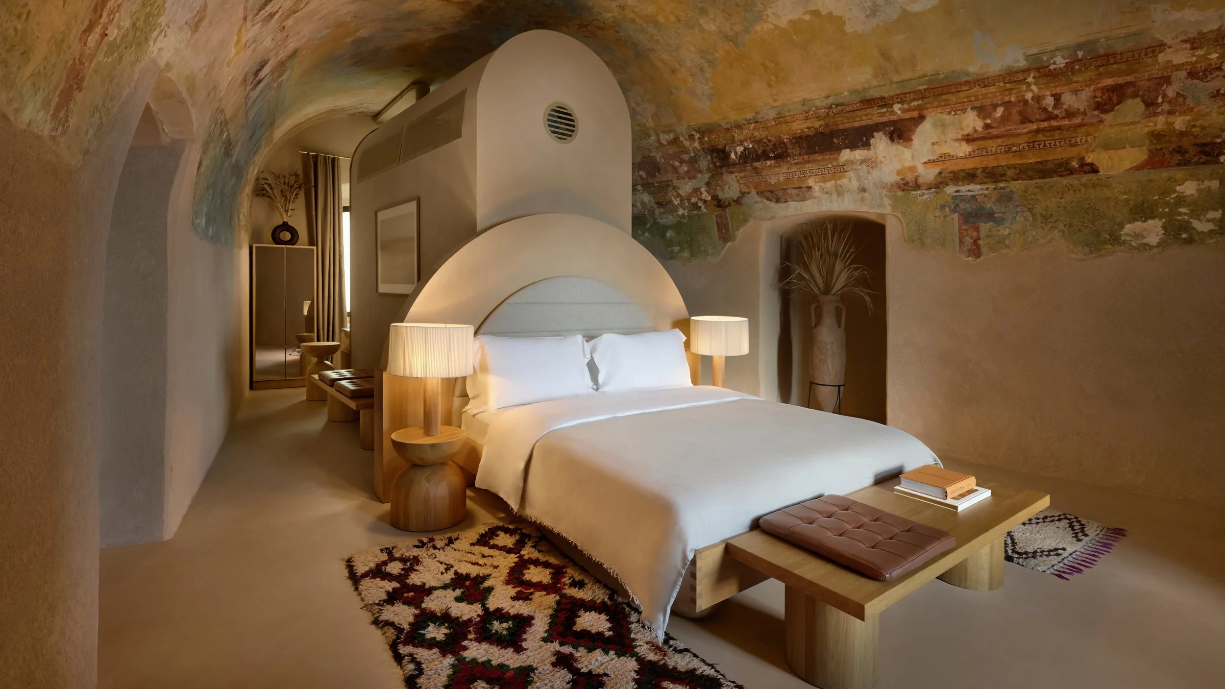 A cozy bedroom with a bed, bedside lamps, a bench, and decorative elements, set in a room with textured, colorful ceiling and walls.