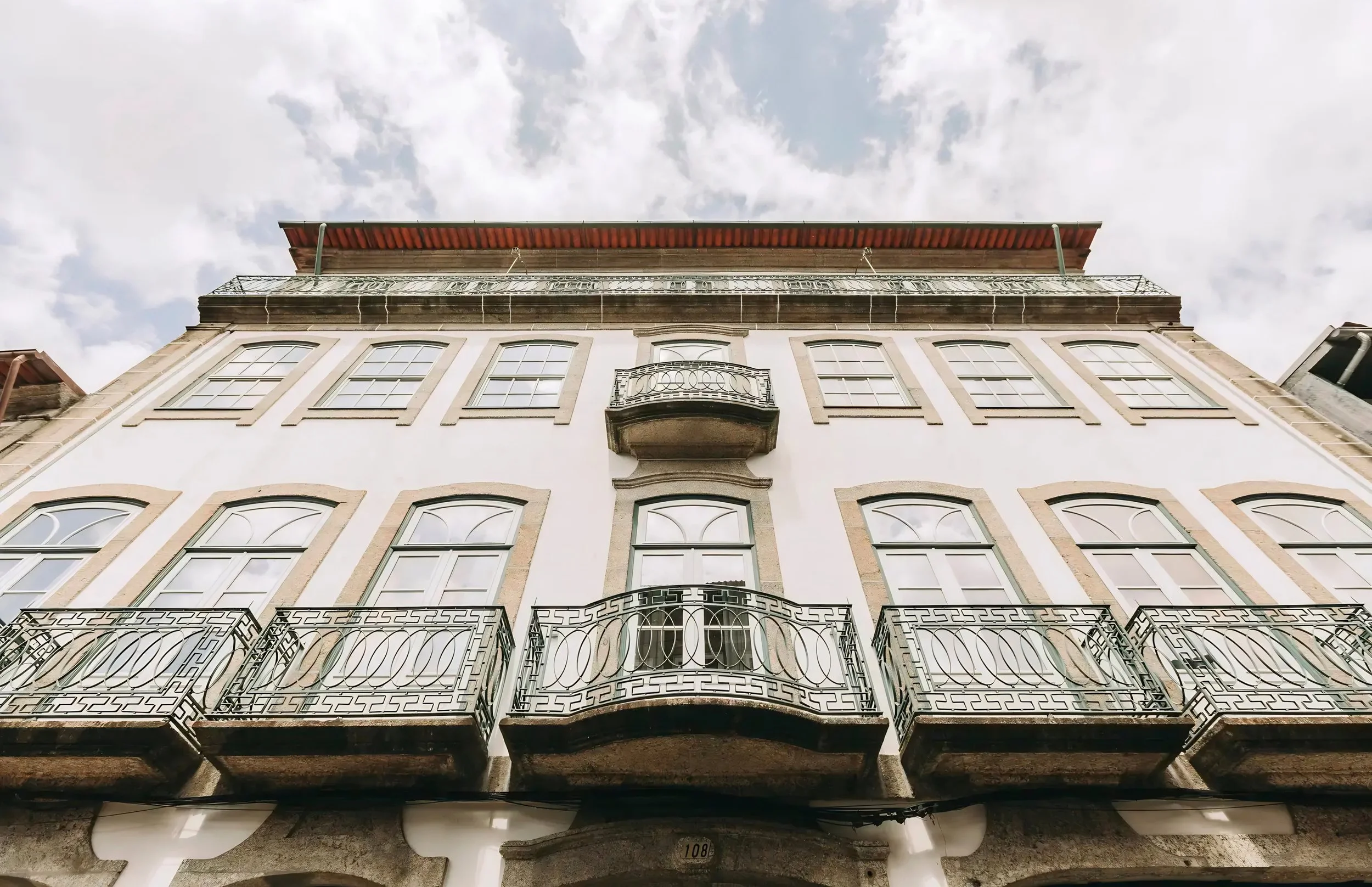 Looking up at a multi-story building with six windows on each level, decorative balcony railings, stone accents around the windows, a small balcony on the third floor, and a red awning on the rooftop, set against a partly cloudy sky.