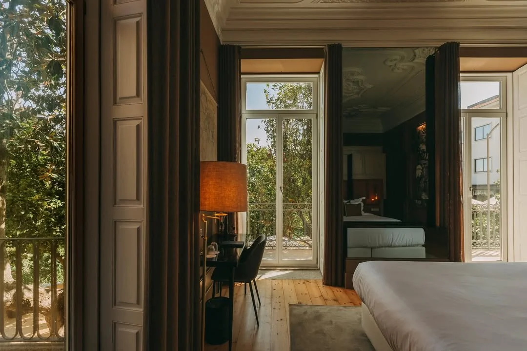 Interior of a hotel room with large windows, a bed, a sitting area, and a balcony with trees outside.