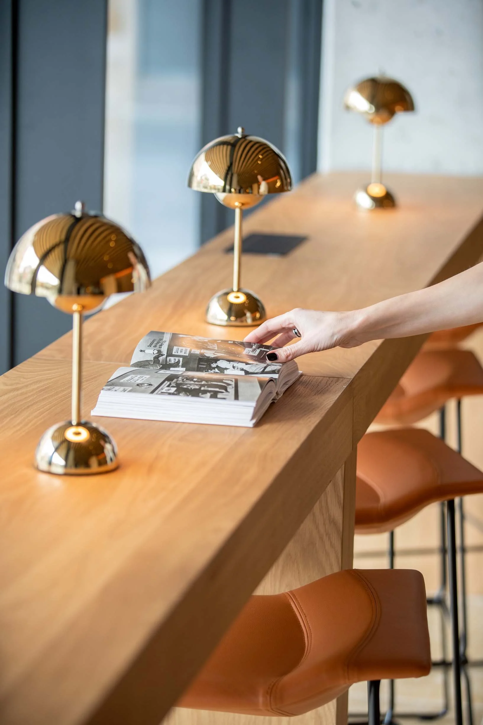 A long wooden table with three modern gold-colored desk lamps and a person with black painted nails turning the pages of a magazine or book on the table. Brown chairs are seen along the side of the table. The setting appears to be a bright, modern interior space.