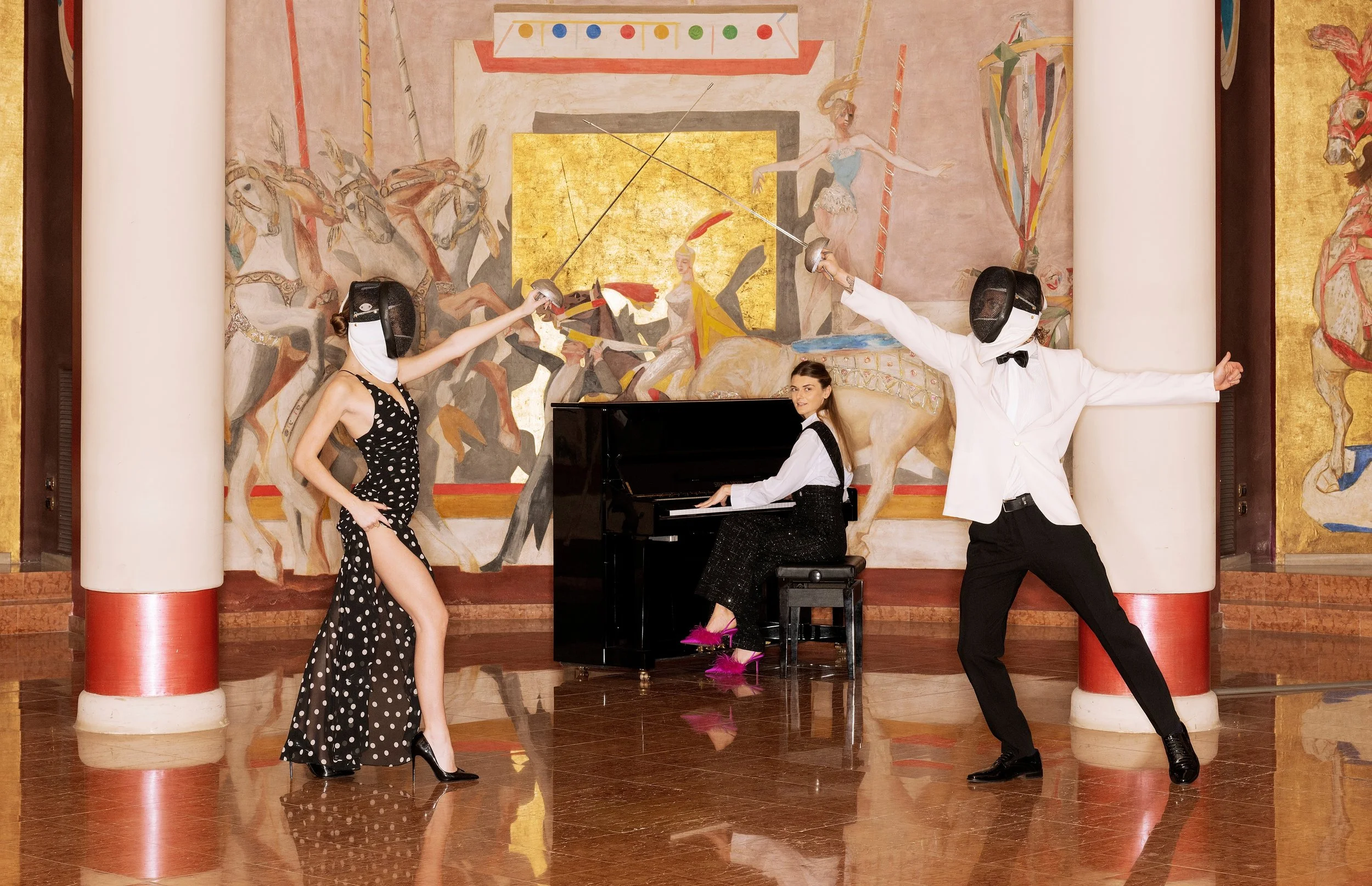 Three people dressed in formal attire, two in fencing masks holding swords, and one woman in a black and white polka dot dress standing in front of a painted mural background. The woman in the polka dot dress is posing with one leg slightly bent, and