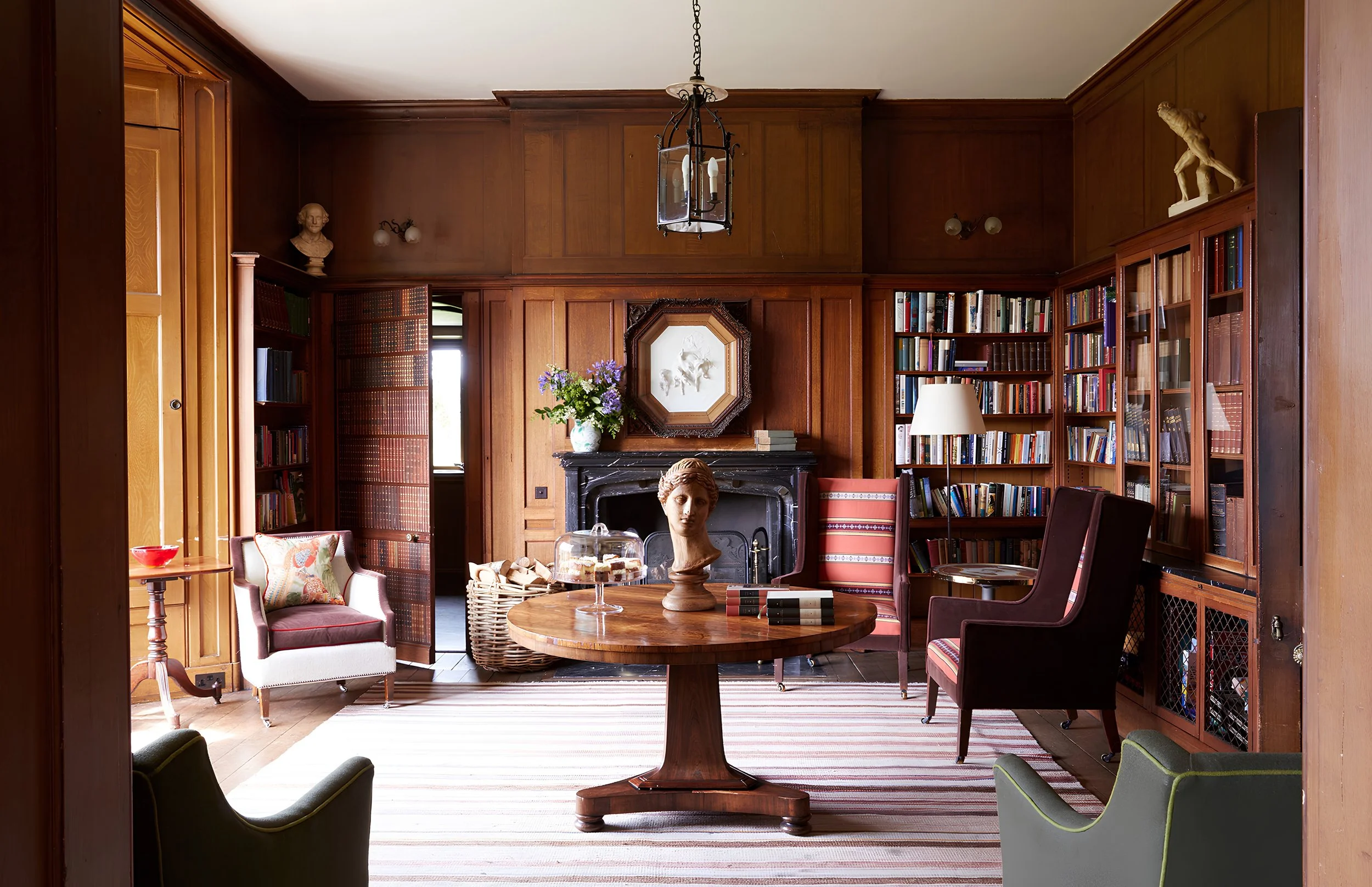 A cozy living room with dark wood paneled walls, a fireplace with a bust sculpture on top, and bookshelves filled with books. There is a round wooden table with a bust sculpture, a cake stand, and a stack of books. The room is decorated with flowers,