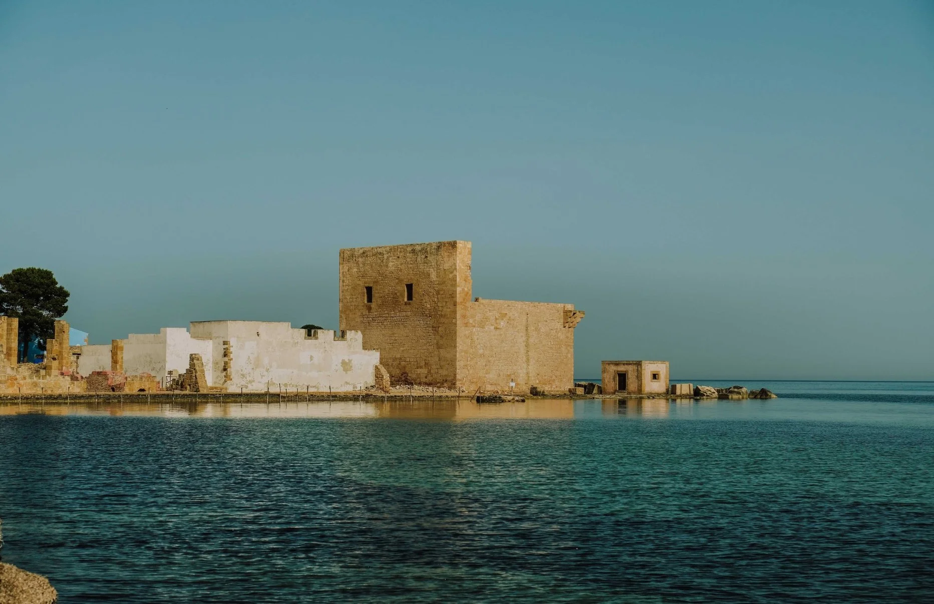 Historical stone buildings and a tower by calm water under a clear blue sky.