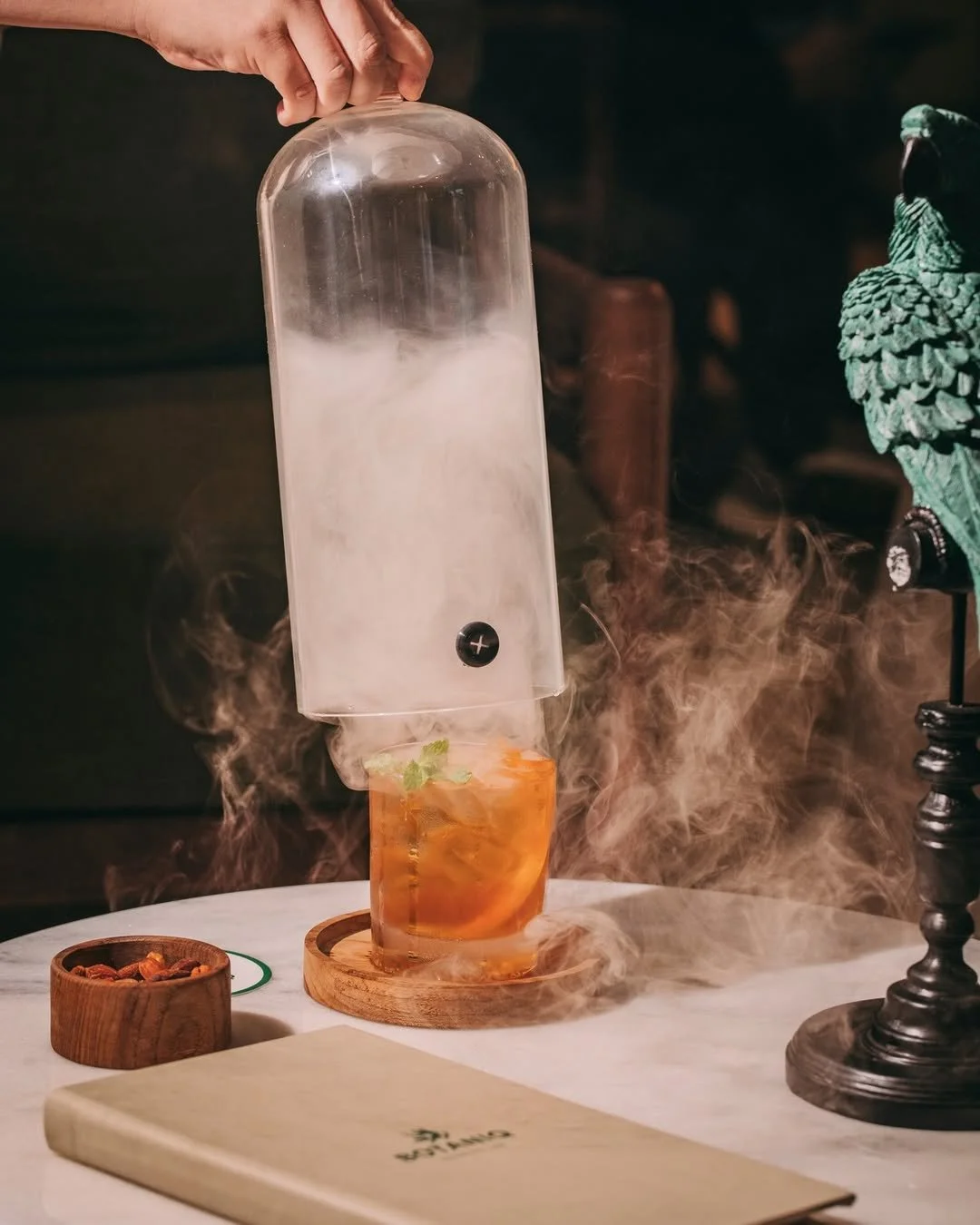 Hand lifting a glass cover over a drink with dry ice fog coming out, on a table with a wooden coaster, a small bowl, a menu, and a decorative statue.
