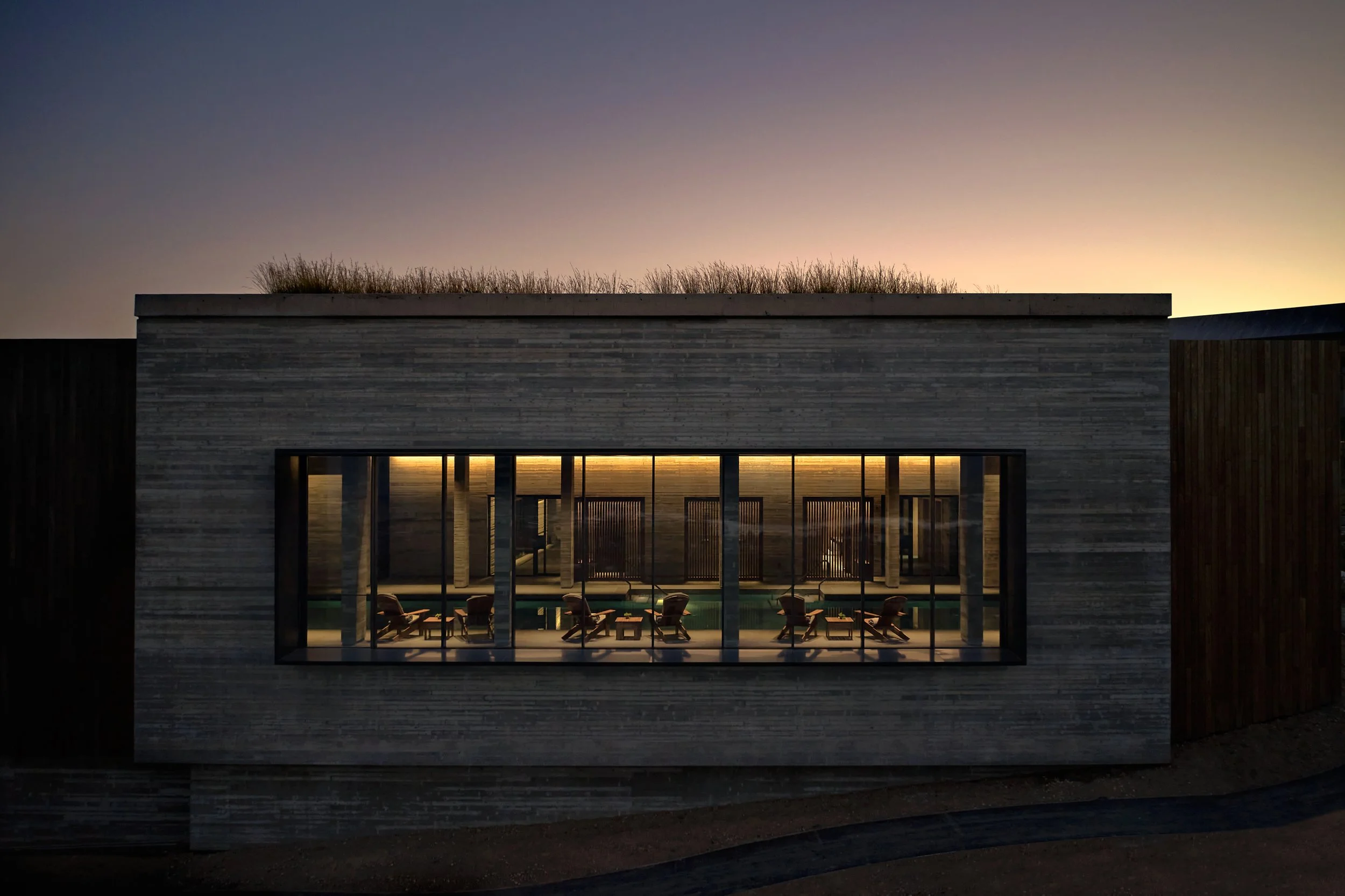 Modern house with large glass windows opening to a patio, illuminated from inside, at dusk.