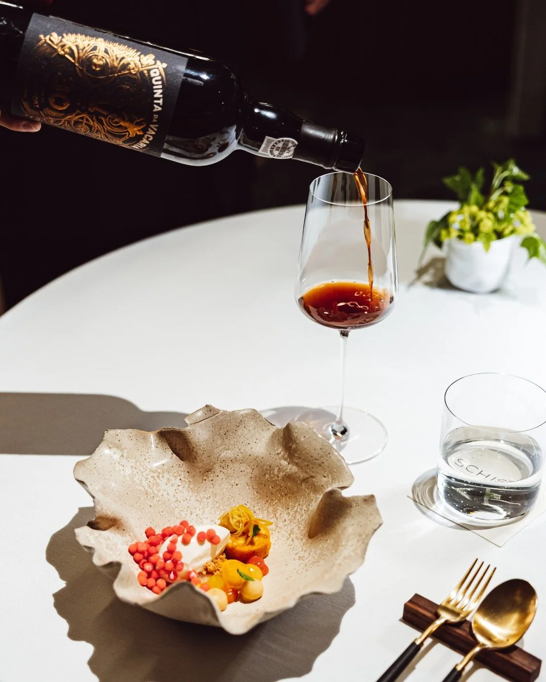 Glass of red wine being poured at a white table with a decorative bowl of desserts, a glass of water, a gold fork and spoon, and a small plant in the background.