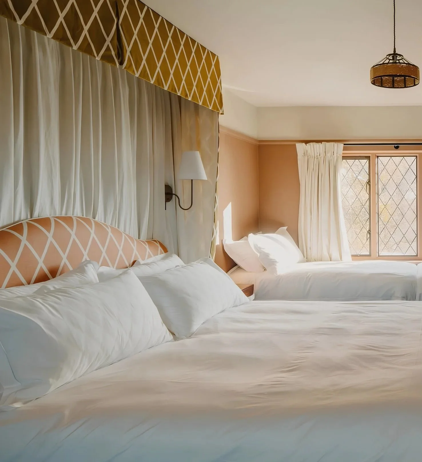 A hotel room with two beds, each with white bedding and pillows. The headboards are patterned with a geometric design. There are curtains over the windows, and a wall sconce lamp between the beds. Natural light illuminates the room.