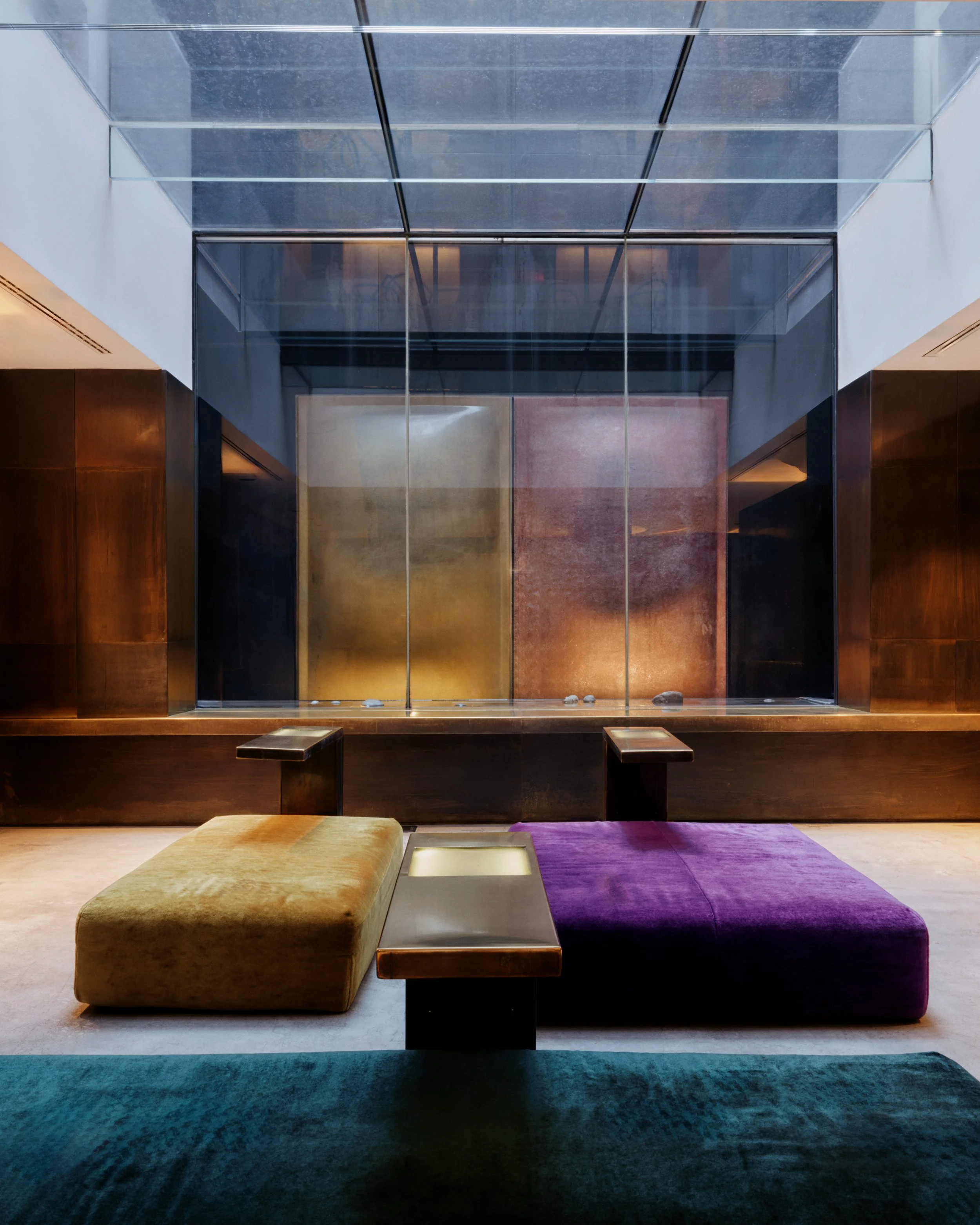 Modern interior with multicolored cushioned seating on a dark carpeted floor, wooden wall panels, and a glass-paneled ceiling with trees visible through it.