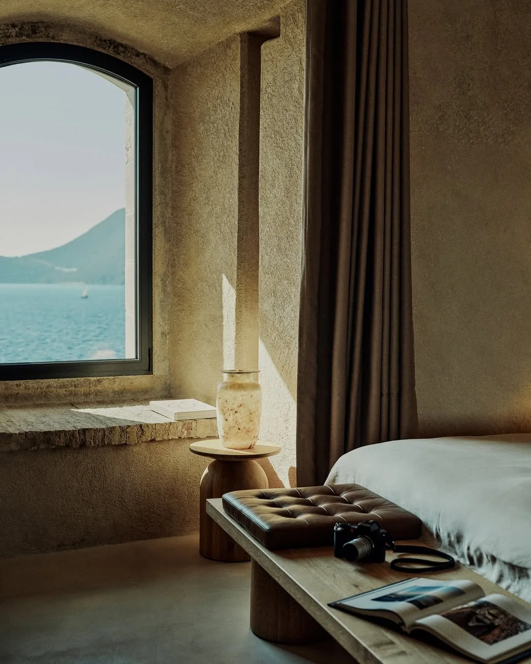 Inside a cozy bedroom with a view of a lake and mountains through a large window, featuring a beige bed, a tufted leather pillow, a wooden bed frame, a camera on the bed, a magazine open, a lampshade, and brown curtains.