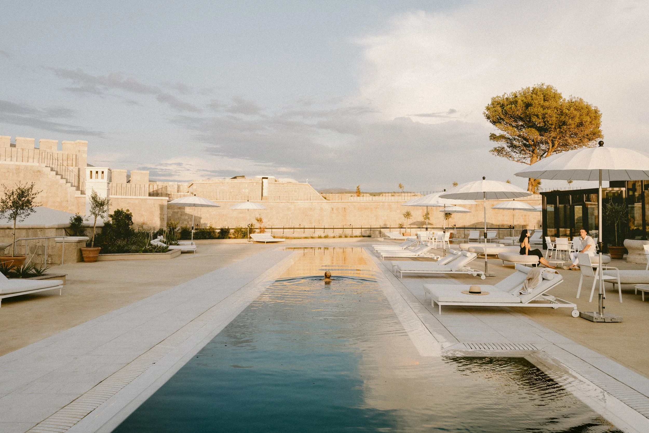 A luxurious outdoor pool area with lounge chairs, white umbrellas, and a couple sitting and chatting under the umbrellas. There are potted plants and a large tree, with a stone wall background and cloudy sky.