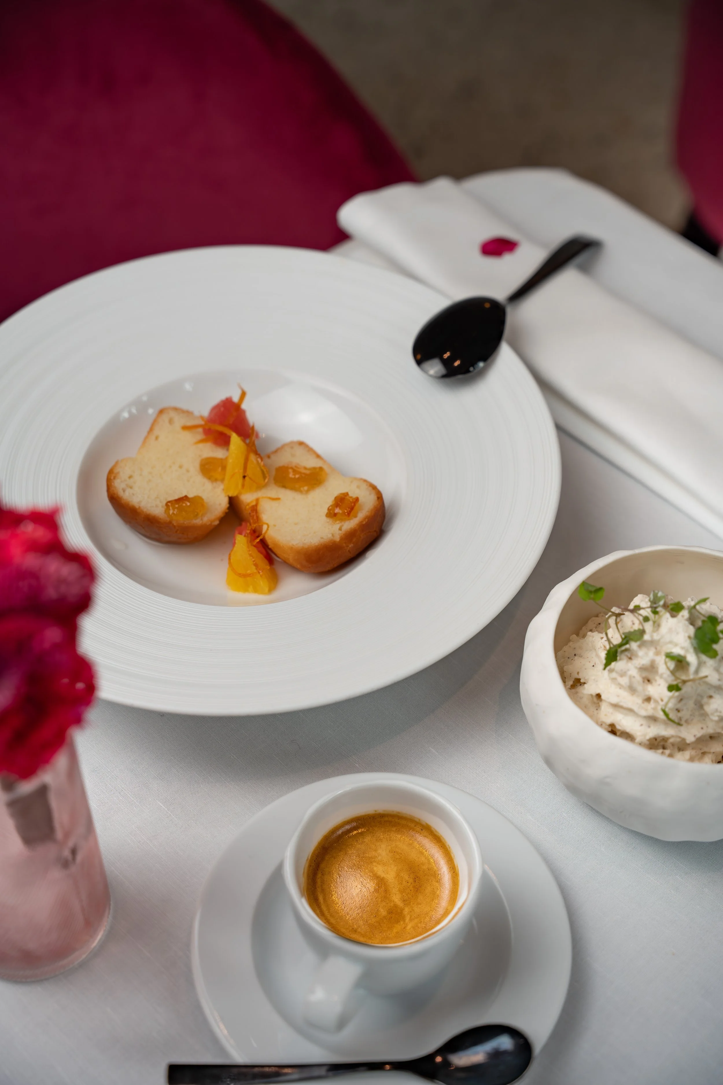 A white coffee cup filled with espresso on a saucer, a white plate with two pieces of cake garnished with yellow and red flower petals, a small bowl of mashed potatoes with herbs, a spoon, and a folded white napkin with a pink flower petal, on a white tablecloth.