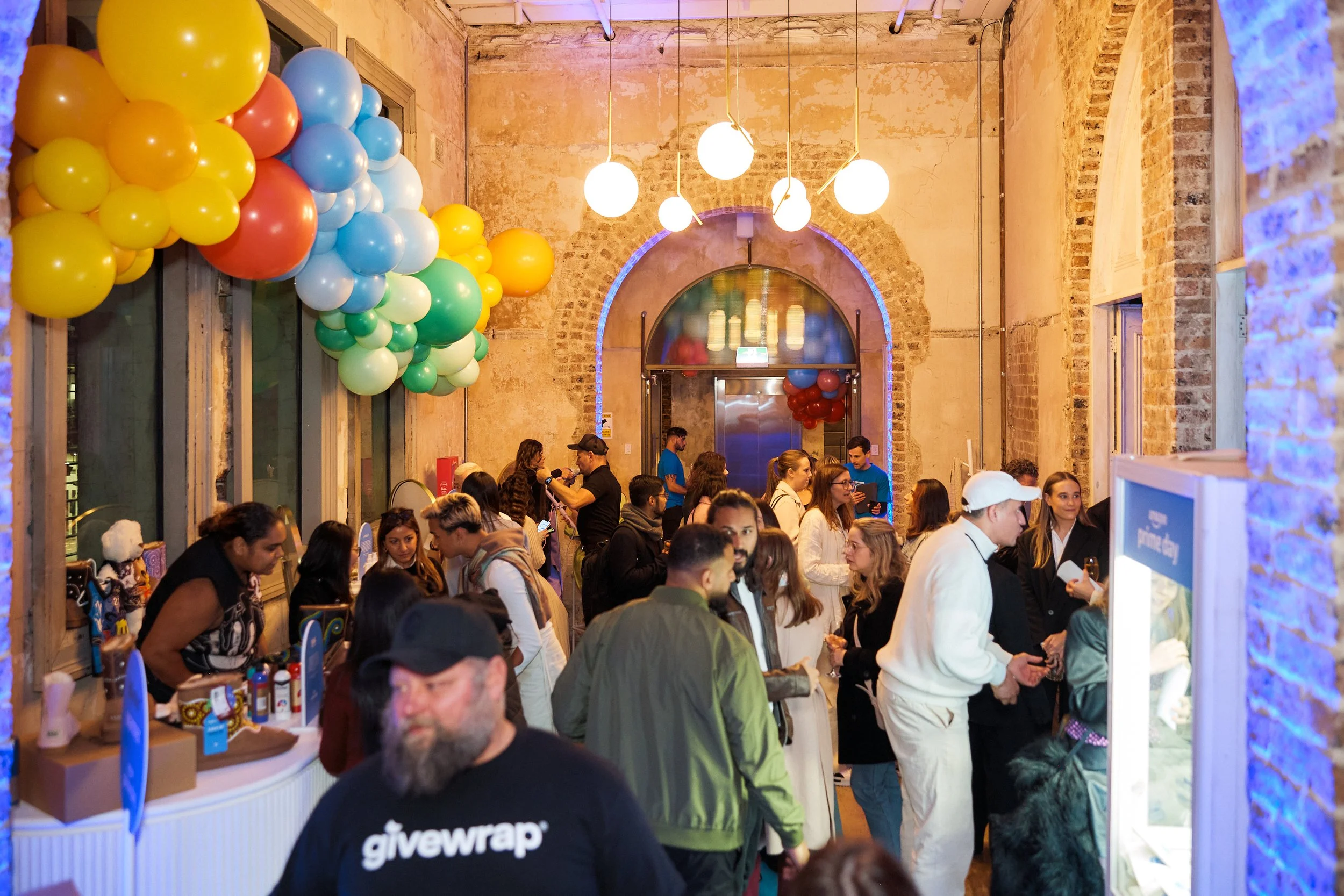 People gather in a decorated indoor space with balloons and hanging lights, possibly at a celebration or event. There are booths with items for sale or display, and the room has brick walls and large windows.