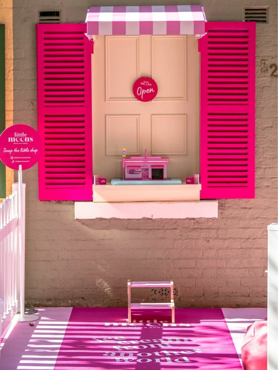 Pink and purple storefront window display with a small toy ice cream shop on a shelf, a pink 'Open' sign, and a pink mat on the ground with white text.