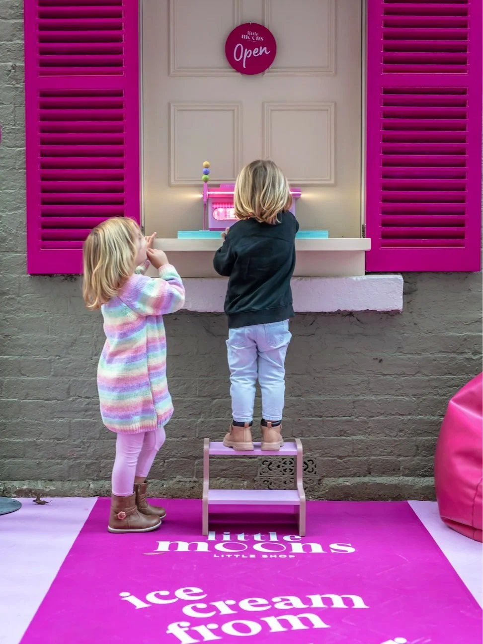 Two young children, a girl and a boy, standing on a pink step stool, looking at a pink and white toy ice cream stand against a beige wall with pink shutters and an 'Open' sign, outside a Little Moons shop.