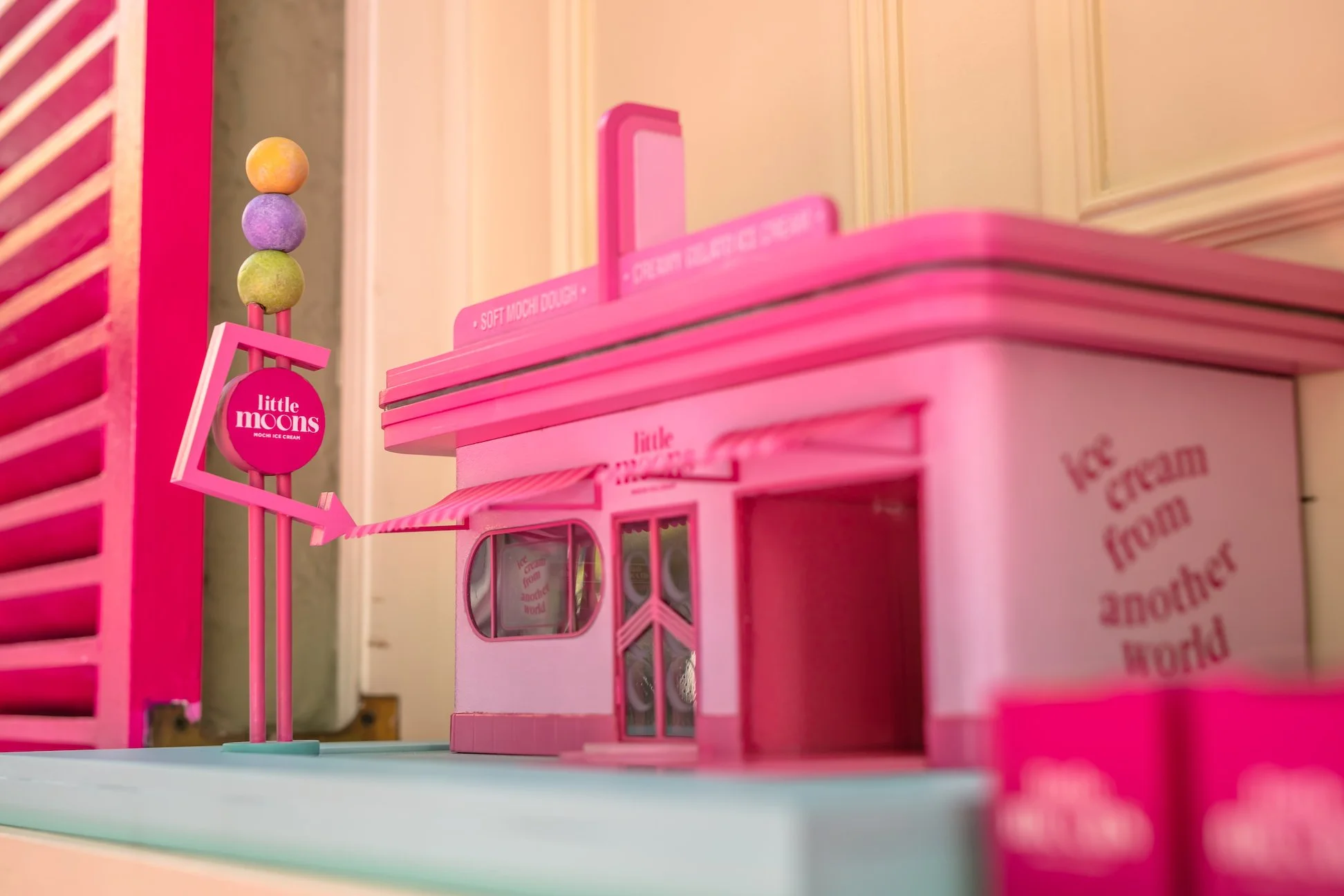 Pink toy shop with a sign reading 'Little Moons' and colorful balls on top, set on a pastel blue surface indoors.