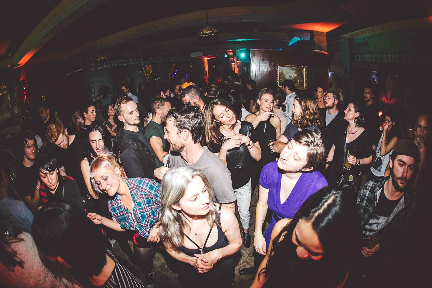 Crowd of people dancing and socializing in a dimly lit nightclub with colorful lighting and paintings on the walls.