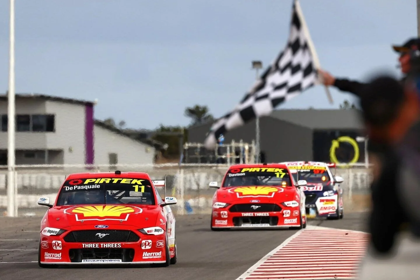 Race cars on a dirt track, leading with a checkered flag being waved by one of the race officials in the background.