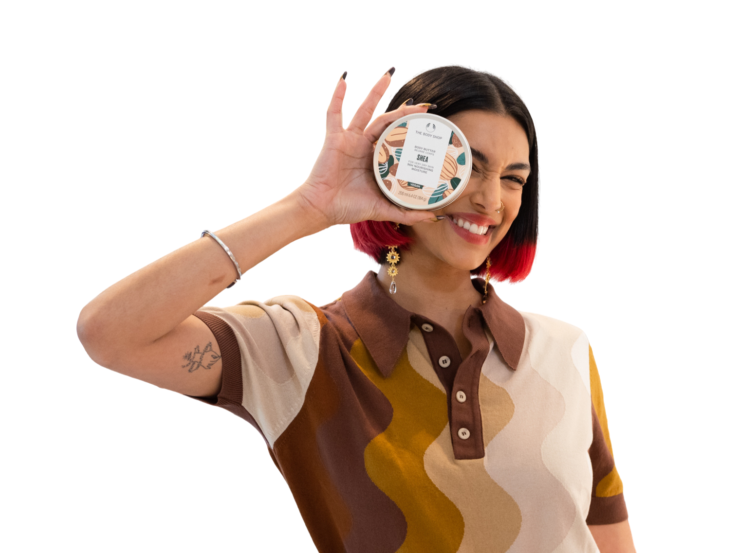 A woman with short black and red hair, earrings, and a tattoo on her arm, smiling and holding a container of body butter near her face.