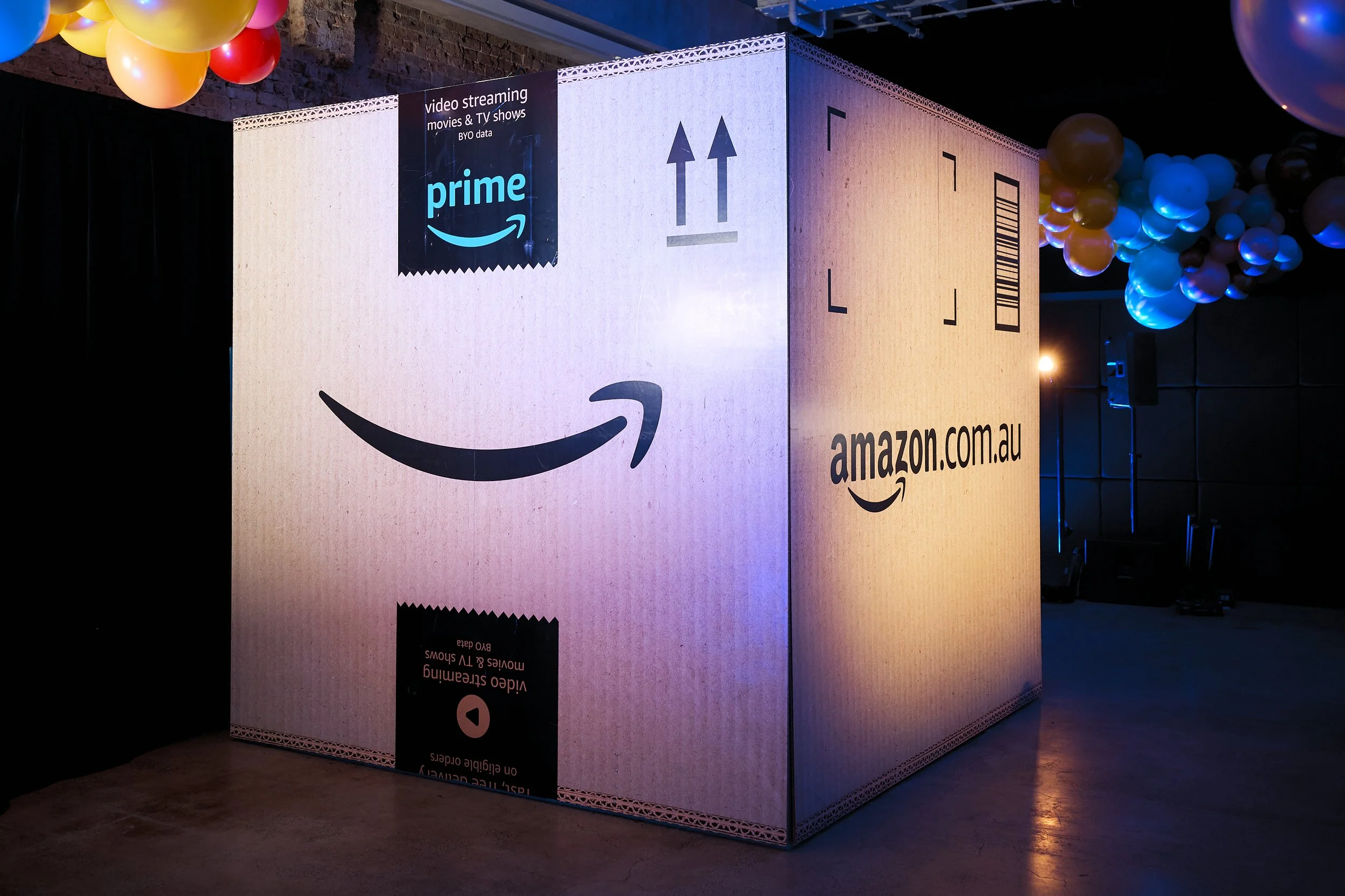 Large cardboard box with Amazon logos, barcode, and arrows, used as a promotional display at an event. The box features the Amazon smiling arrow, references to Prime video streaming and the Australian website amazon.com.au. Balloons are seen overhead