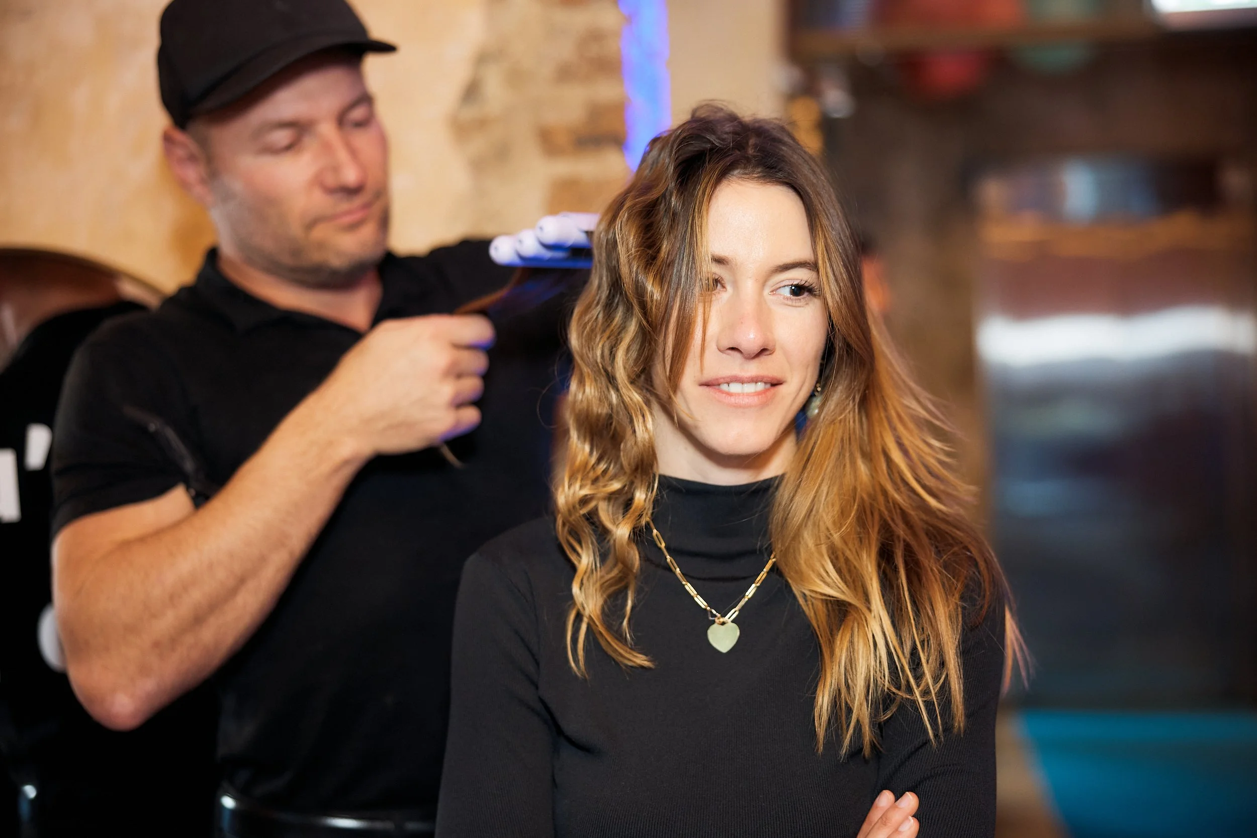A hairdresser is blow-drying a woman's hair in a salon. The woman is smiling and wearing a black top and a heart-shaped pendant necklace.