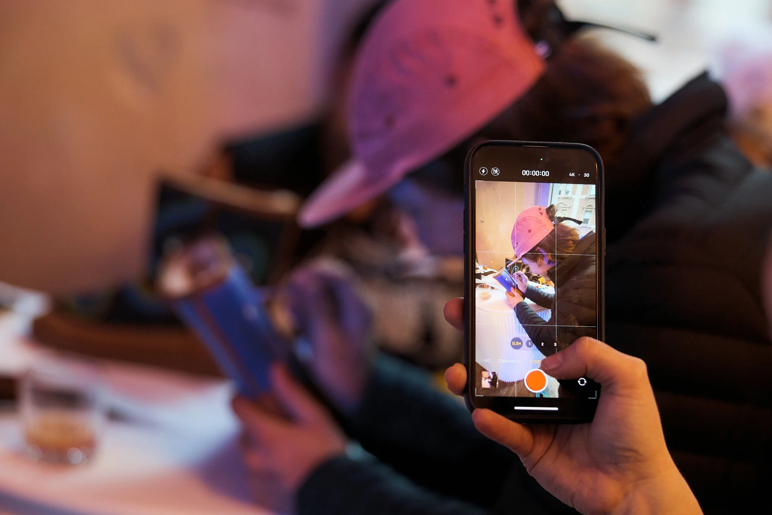 Person filming a woman wearing a pink baseball cap using a smartphone at a table in a cafe or restaurant.