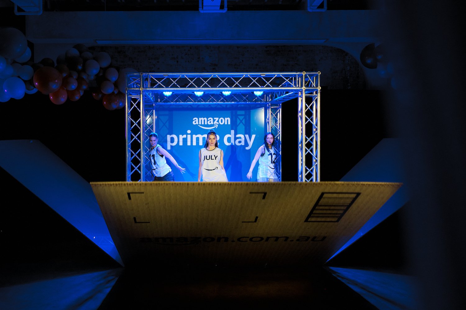 Stage with three girls performing at an Amazon Prime Day event, with a large screen behind them displaying the Amazon Prime Day logo, in a dark indoor setting.