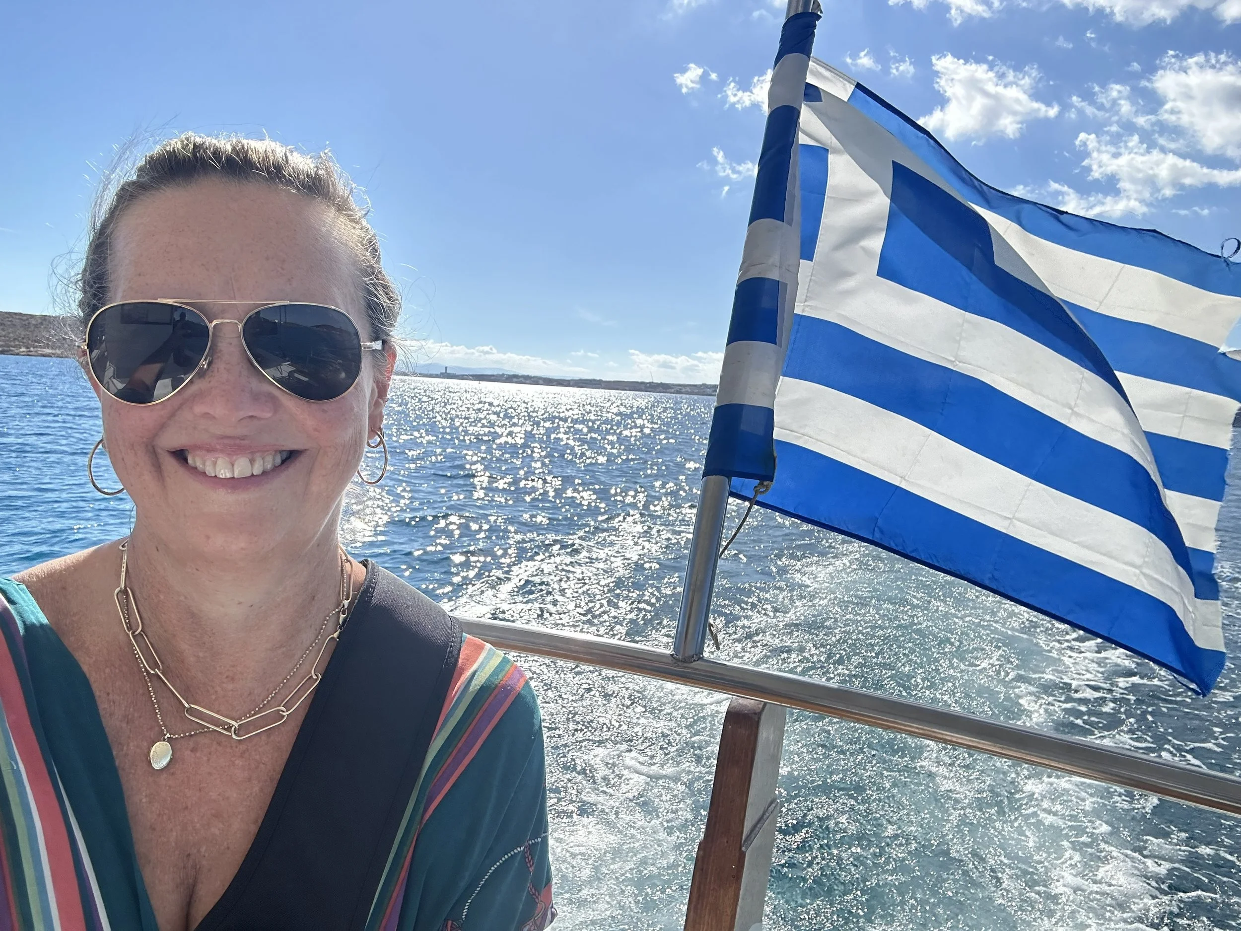 A woman smiling while taking a selfie on a boat. She is wearing sunglasses, necklaces, and a colorful striped shirt with a dark vest. Behind her, a blue and white flag waves in the wind, with a body of water and a partly cloudy sky in the background.