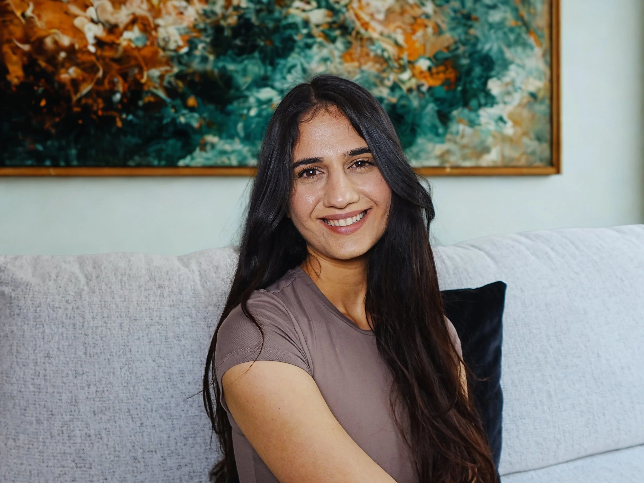 A woman with long dark hair sitting on a light-colored couch, smiling at the camera. There is an abstract painting with green, orange, and white colors hanging on the wall behind her.