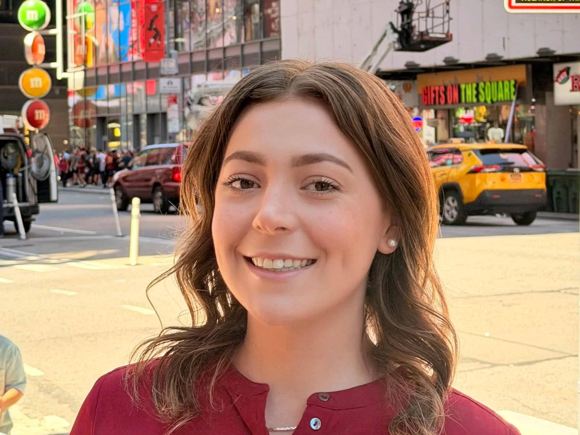 Close-up of a young woman with long brown hair smiling outdoors during daytime, with trees, bicycles, and a statue in the background.