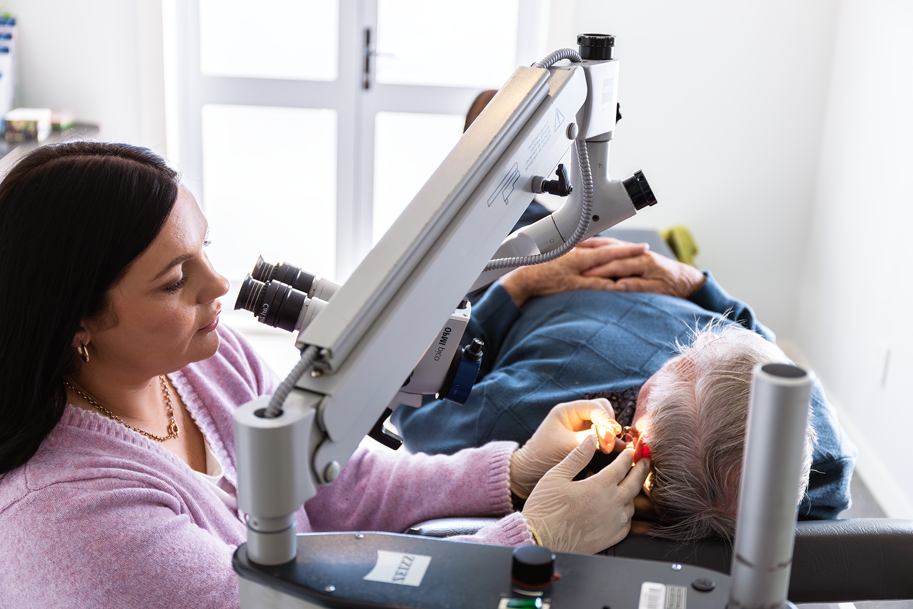 A woman performing eye surgery on an elderly patient with a medical microscope in a clinical setting.