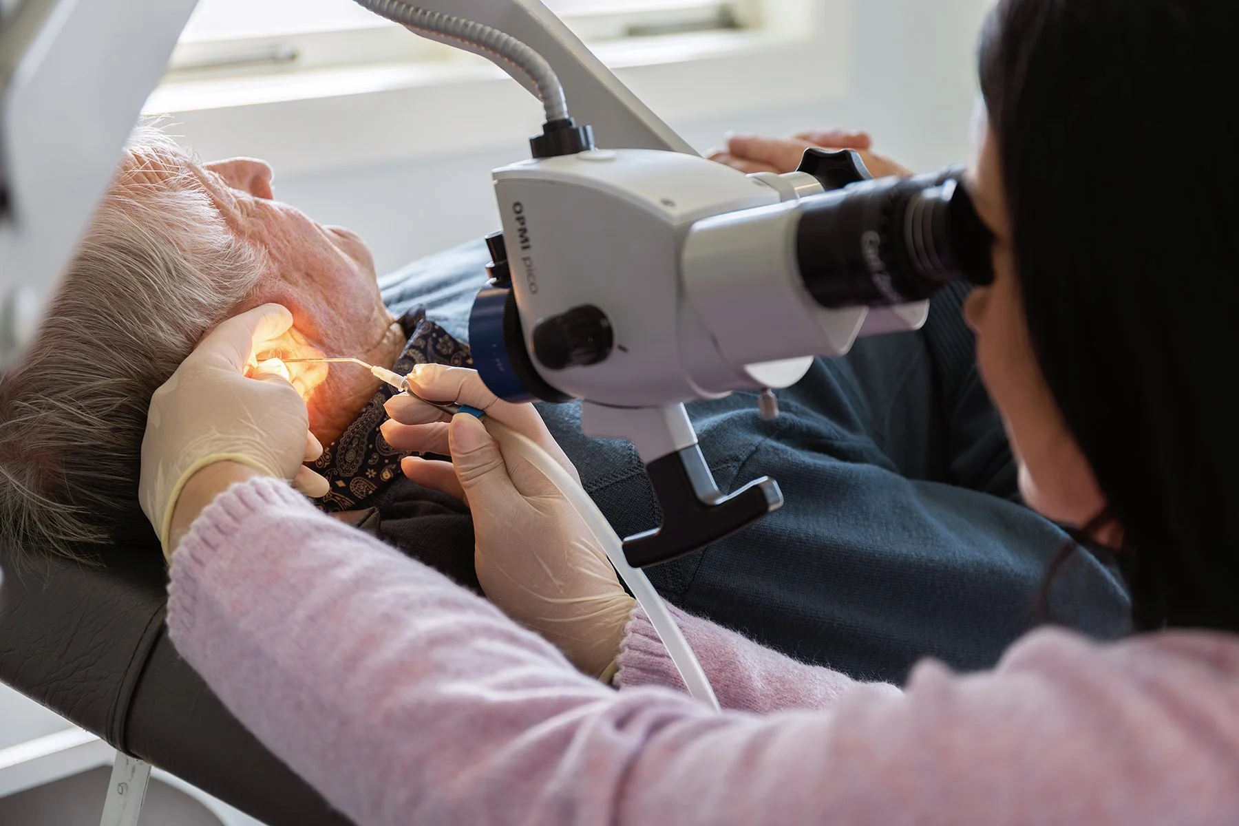 A senior woman lying in a medical chair getting an ear examination with a doctor using an otoscope and a surgical light.