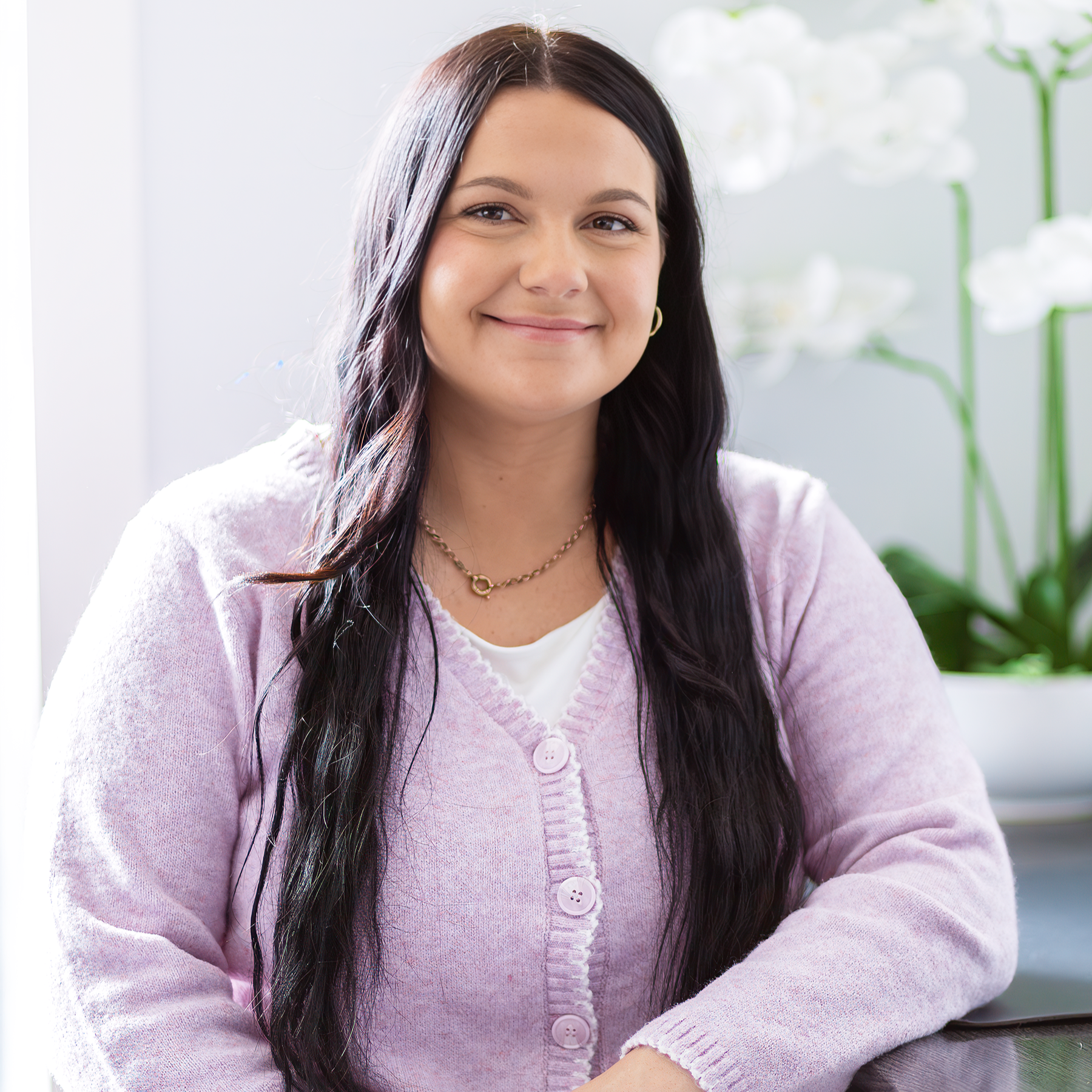 A young woman with long dark hair sitting indoors near a window with white flowers in the background, wearing a light pink cardigan and smiling at the camera.