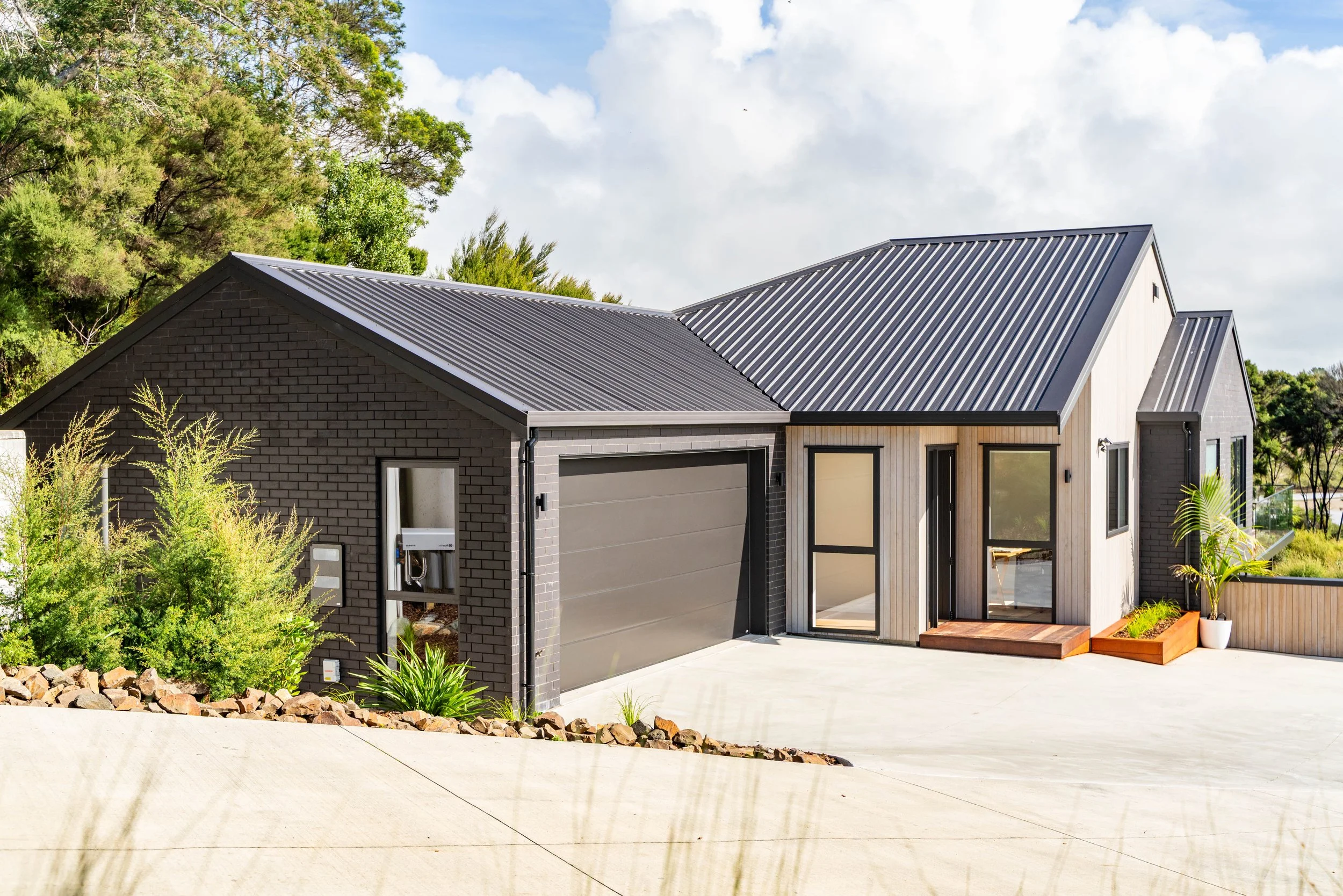 Modern house with black brick and light-colored wood siding, metal roof, large windows, and a garage, surrounded by greenery and a concrete driveway.