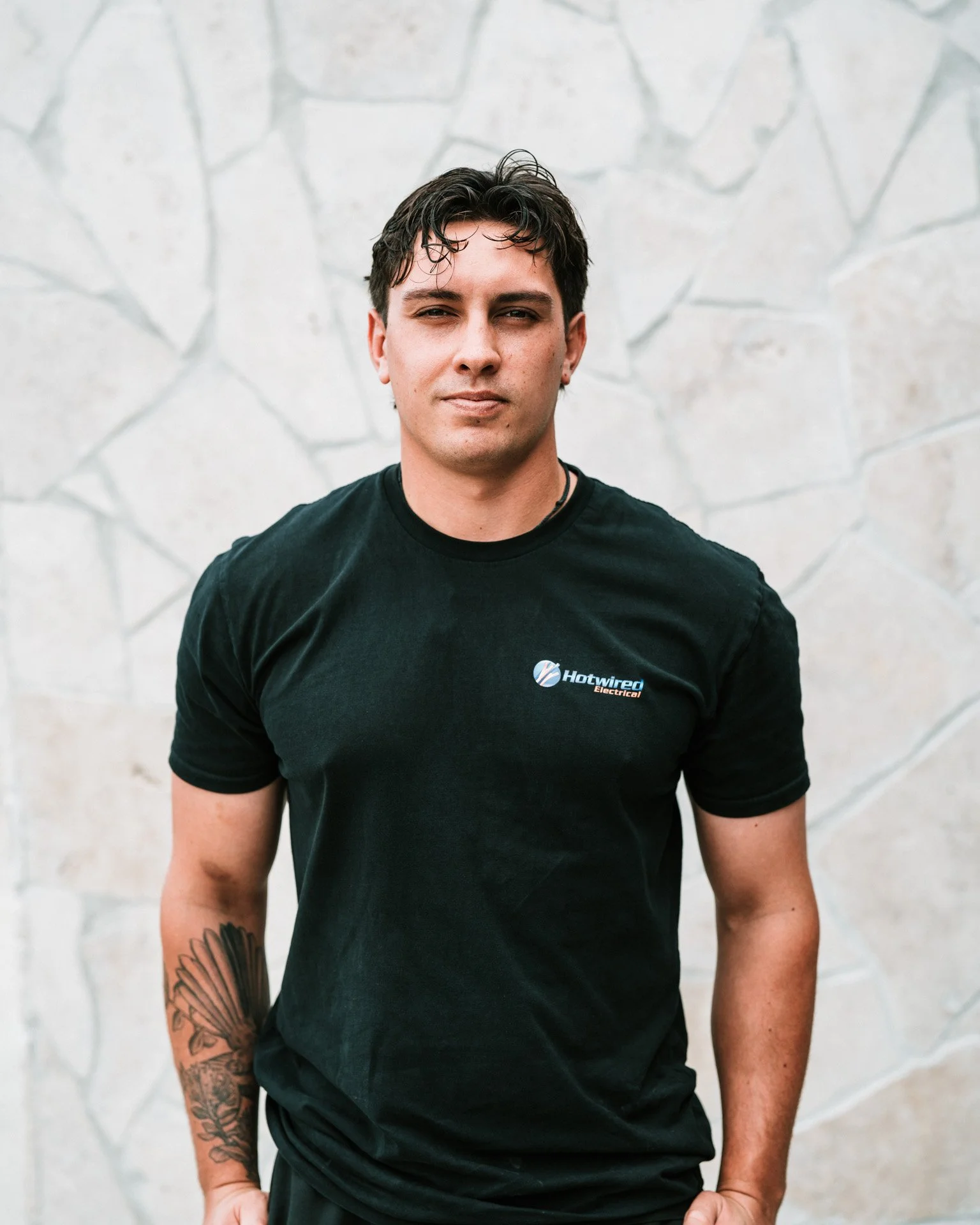 A young man with dark hair and a tattoo on his left arm, wearing a black T-shirt with a logo that says 'Hotwired Electrical,' standing against a stone wall.