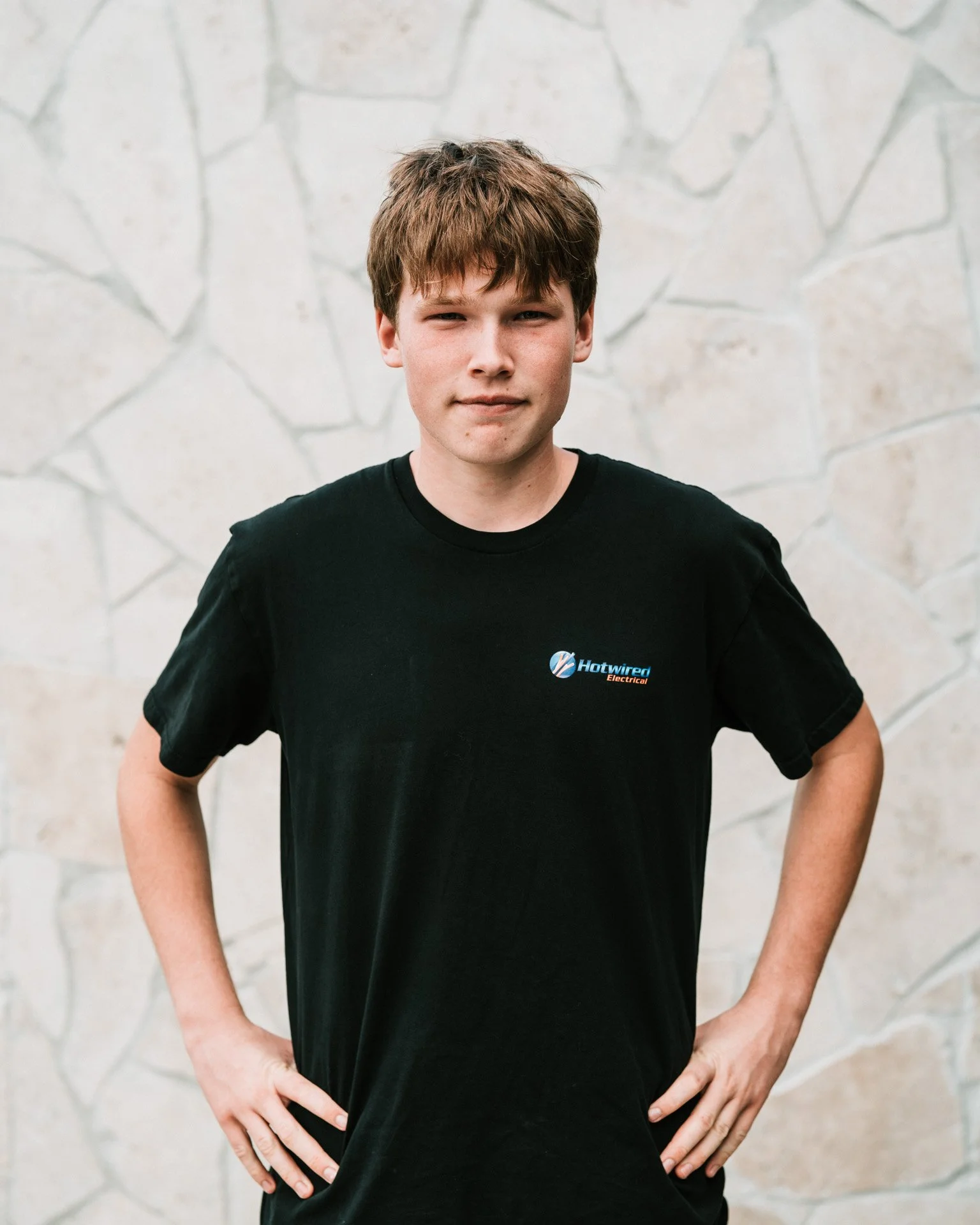 A young man with brown hair and fair skin standing outdoors against a stone wall, wearing a black t-shirt with a small logo on the chest.
